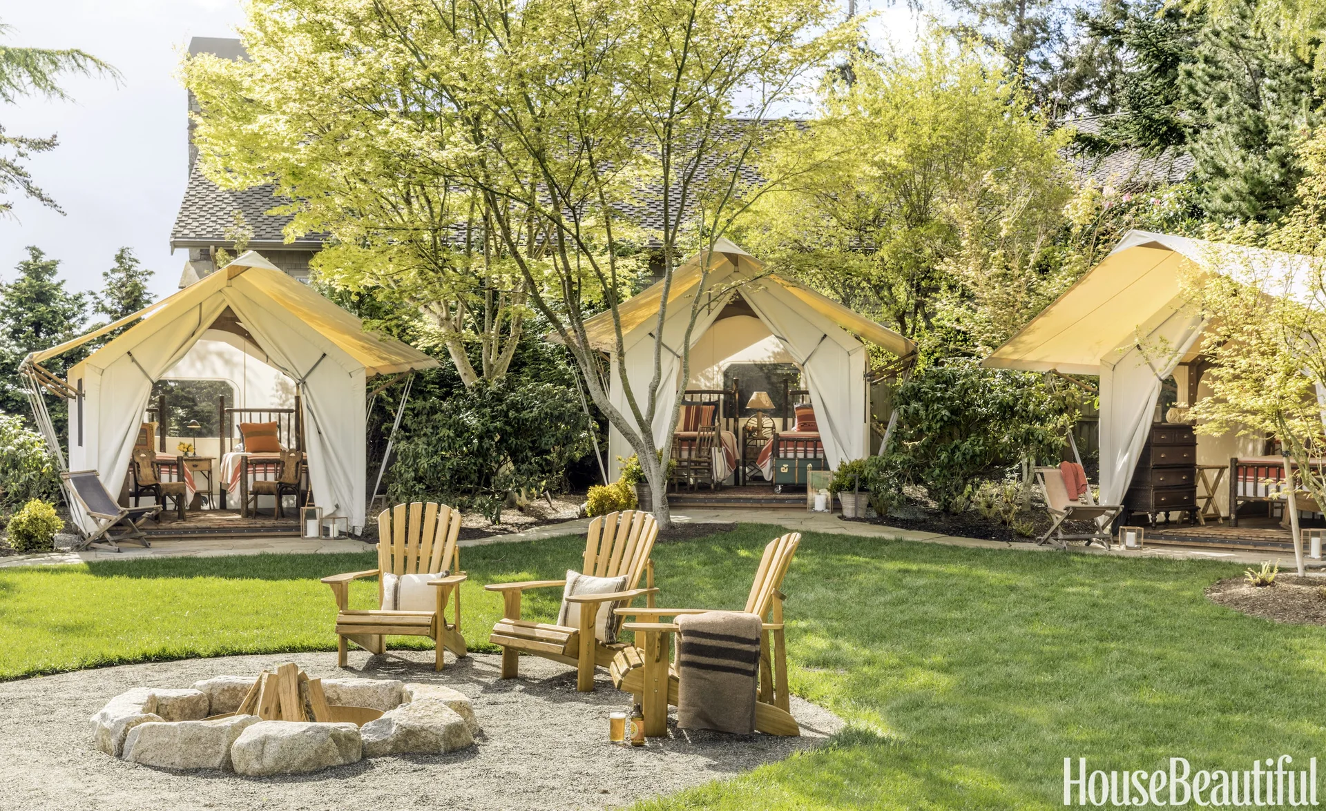 Three canvas glamping tents stand behind a stone-ringed fire pit surrounded by wooden Adirondack chairs on a grassy lawn.