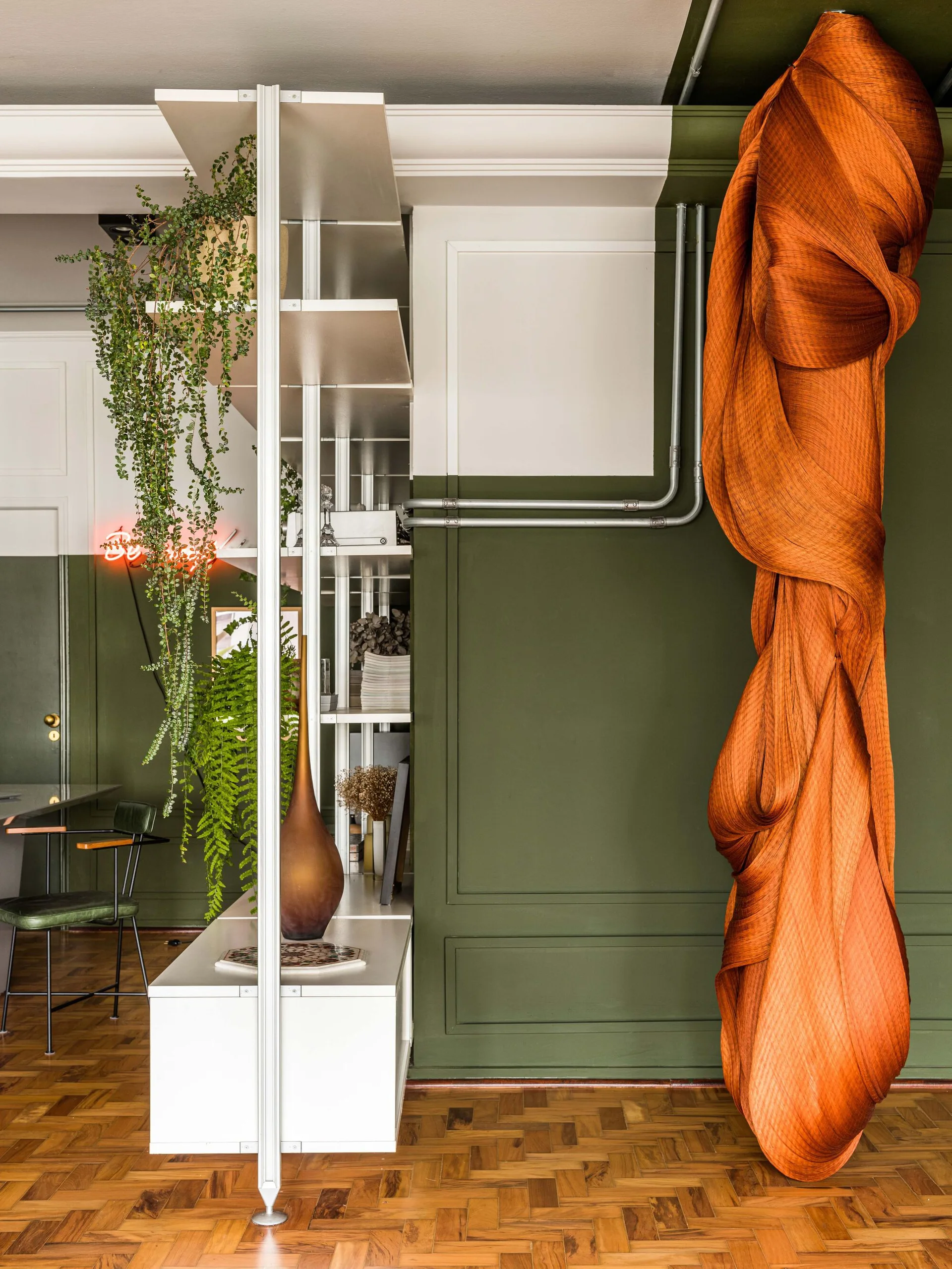 An olive green room features a white metal open shelving unit next to a large draped orange fabric sculpture on parquet floors.