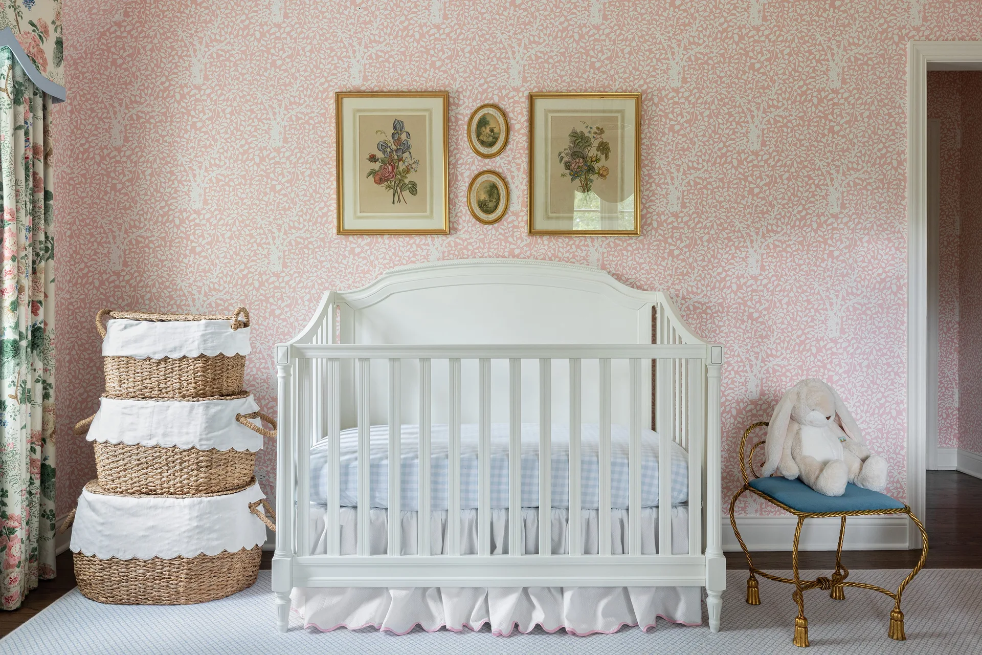 A white wooden crib sits against pink floral wallpaper, next to stacked woven baskets and a gold stool with a plush toy.