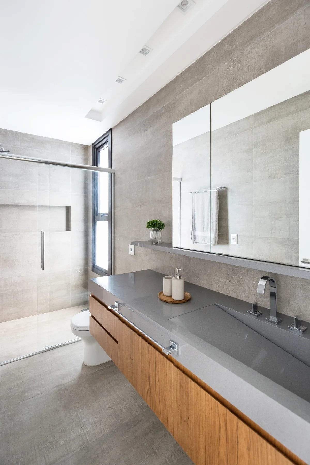 A modern bathroom featuring a floating wooden vanity with a grey stone-like countertop, matching wall tiles, and a glass shower enclosure.