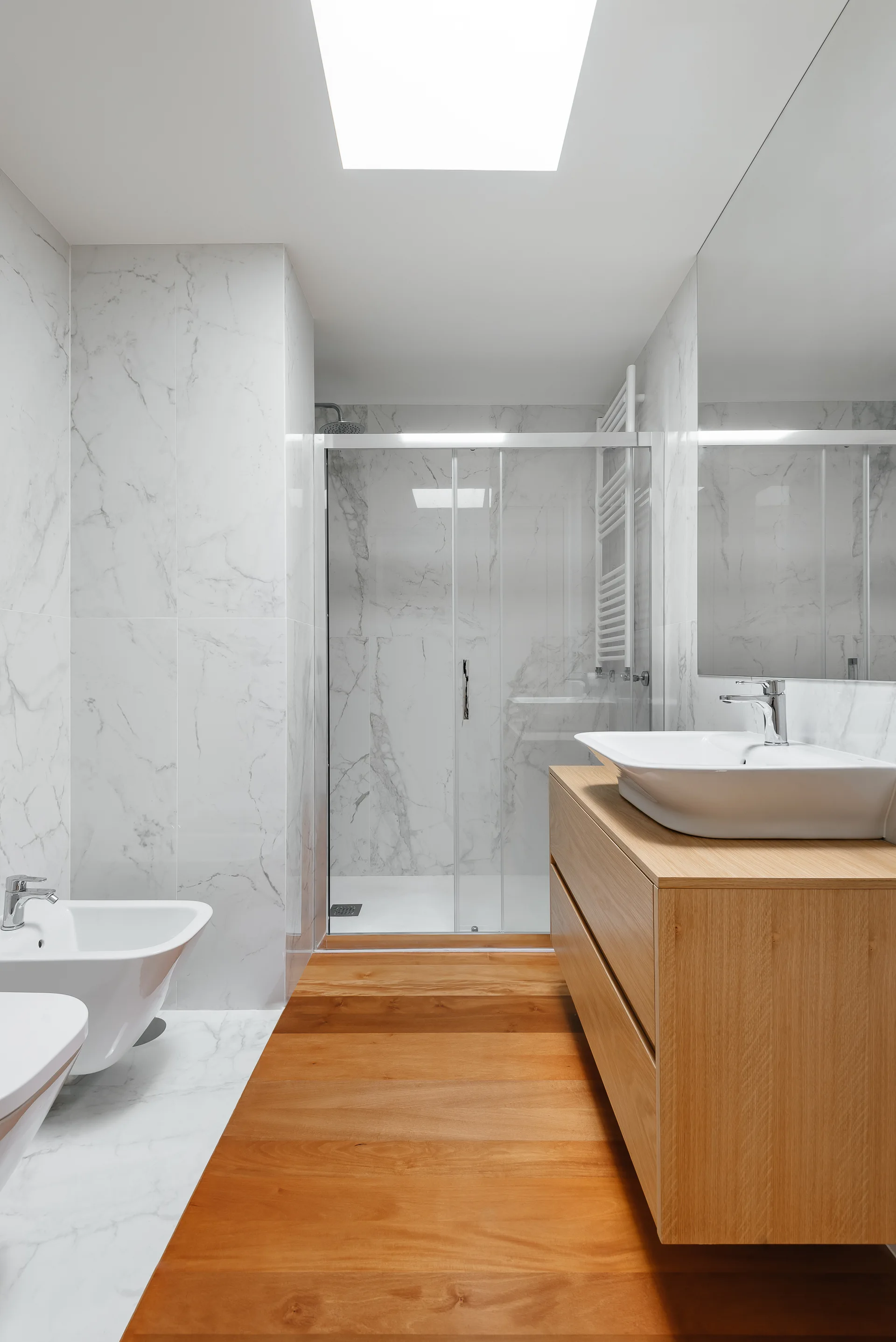 A modern bathroom featuring marble tiled walls, a floating wood vanity with a white vessel sink, a glass-enclosed shower, and warm wood flooring.
