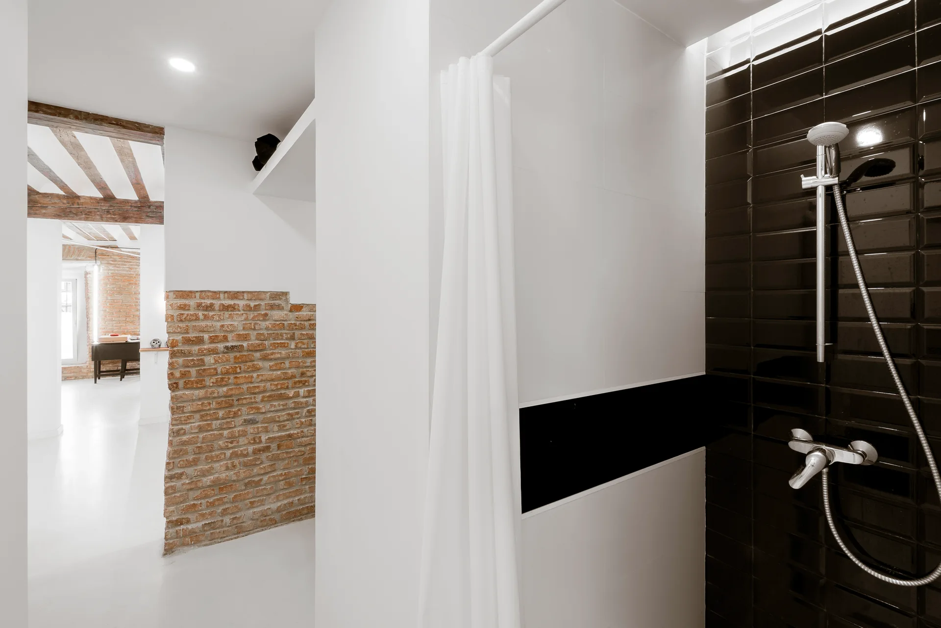 A bathroom featuring a glossy black subway tiled shower with chrome fixtures next to a white wall and a rustic brick wall in the background.