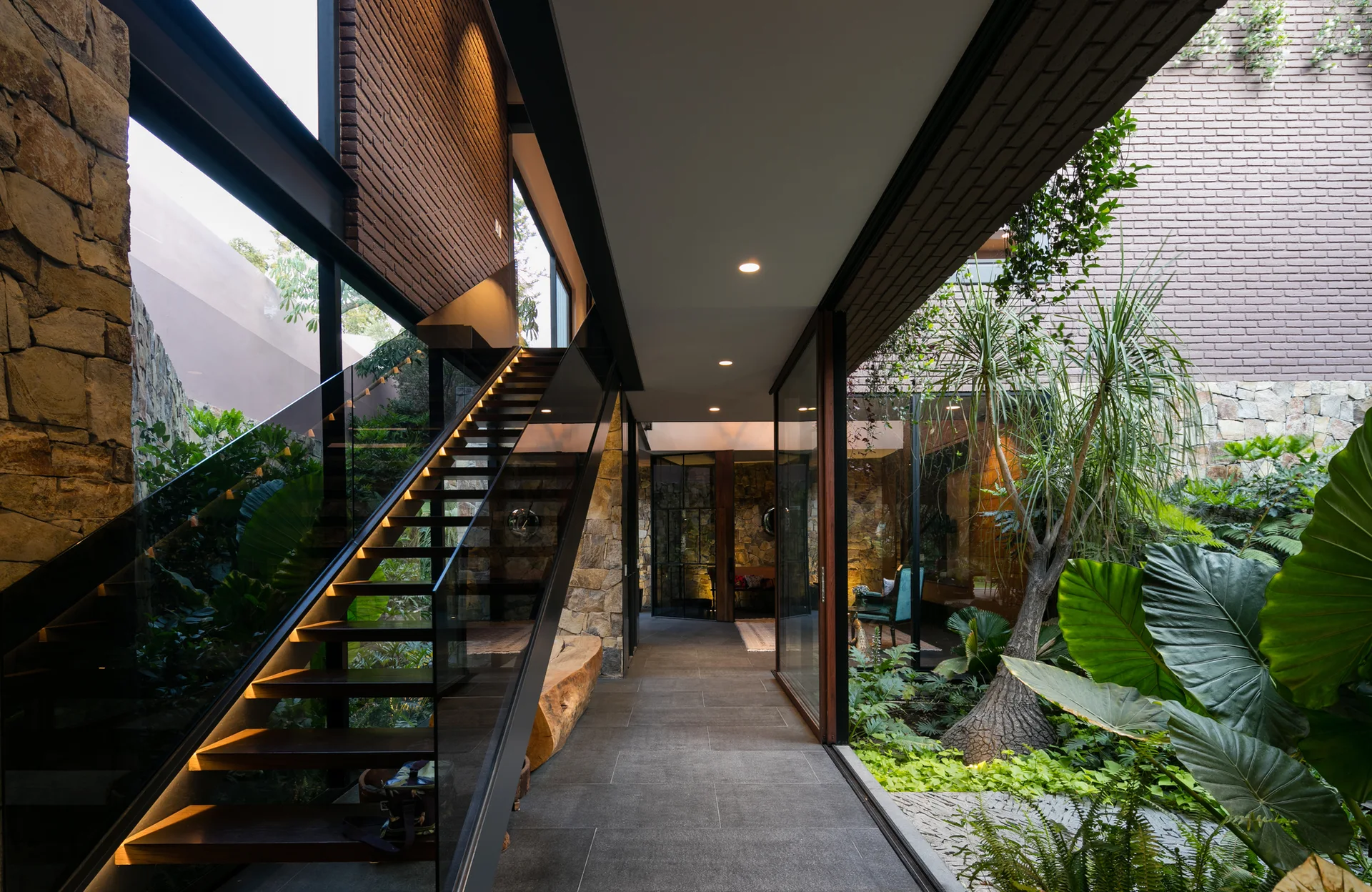 A modern tiled hallway features a floating wooden staircase with glass railings alongside a glass-walled courtyard filled with lush green plants.