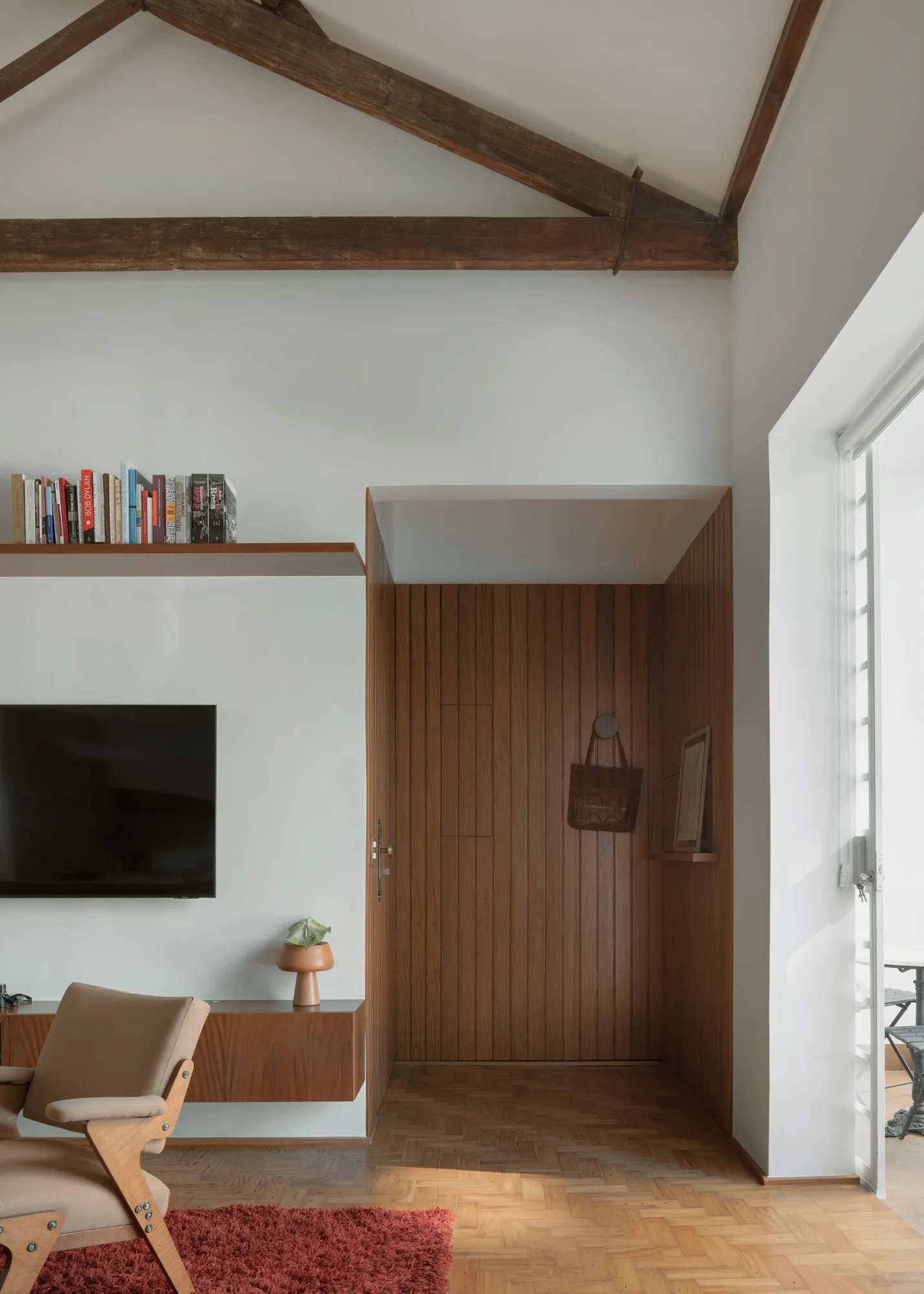 A living room features a wood-paneled entryway, exposed ceiling beams, a floating wooden media console, and a red textured rug.
