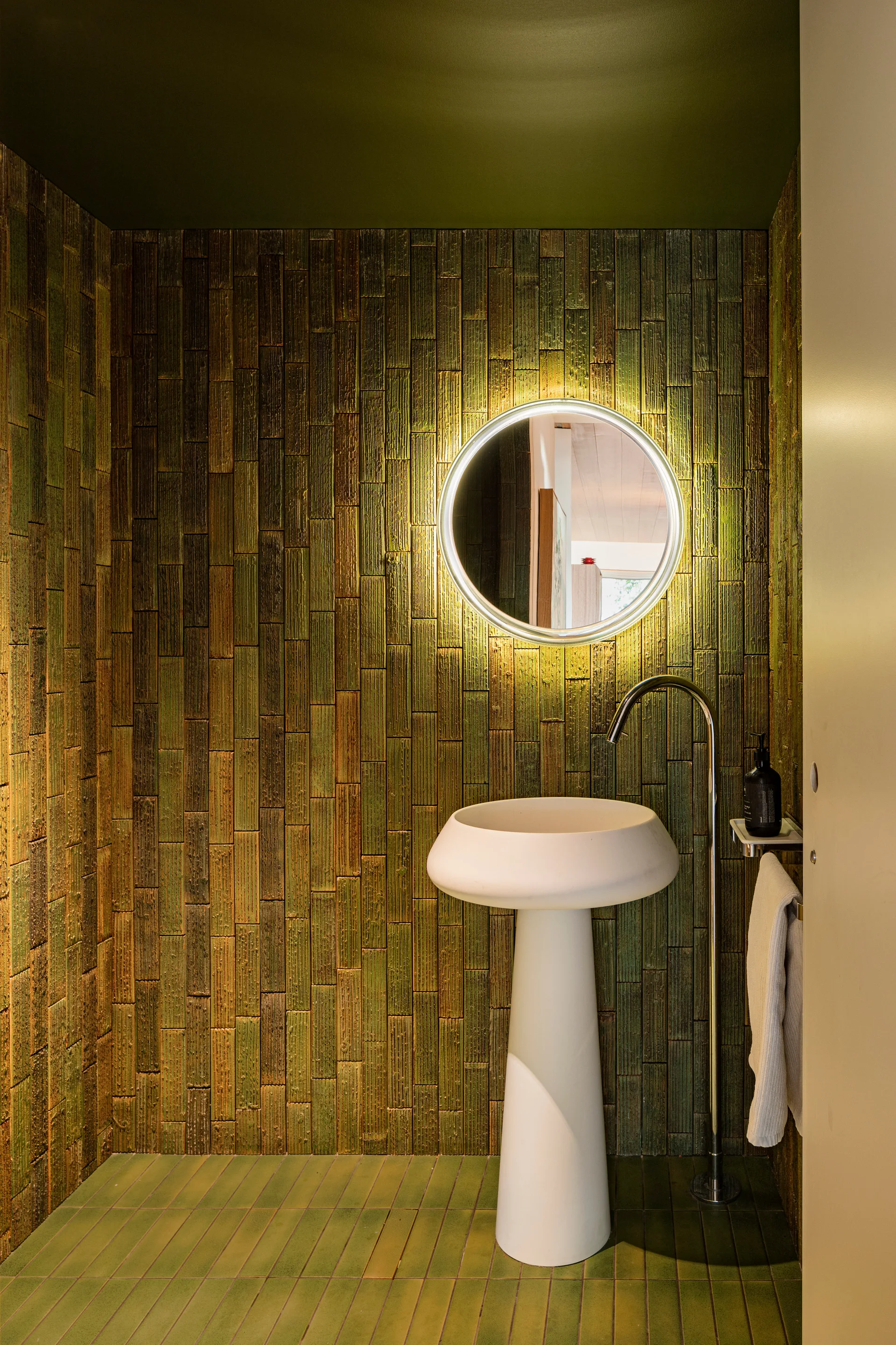 A compact powder room with textured green vertical wall tiles, a sculptural white pedestal sink, and a circular backlit mirror.