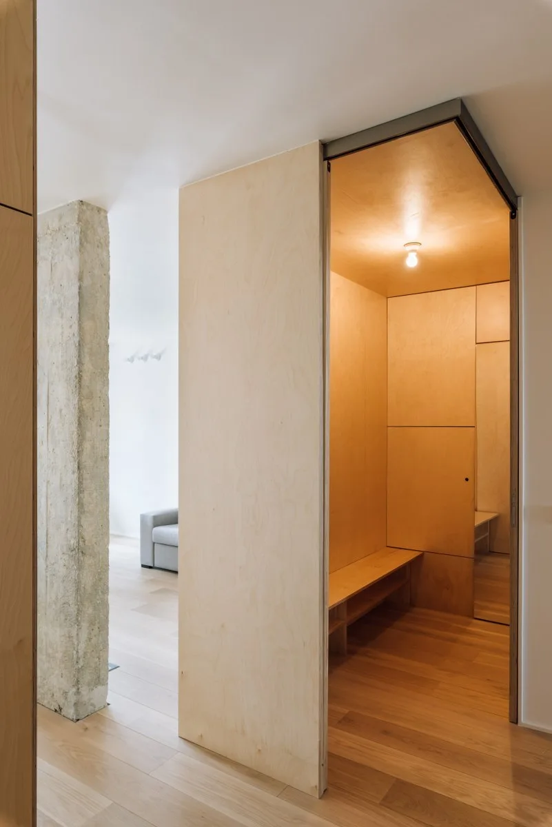A light-wood plywood entryway alcove features a built-in bench, situated next to a raw concrete pillar in a minimalist home.