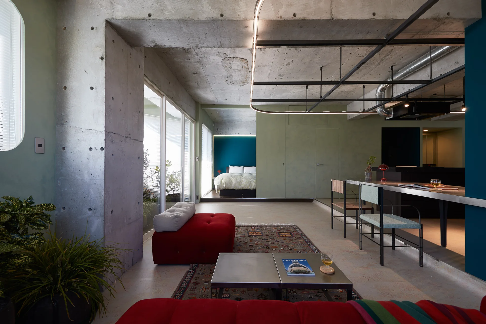 An open industrial living room featuring raw concrete pillars, a bold red sofa, a low metal coffee table, and a bedroom in the background.