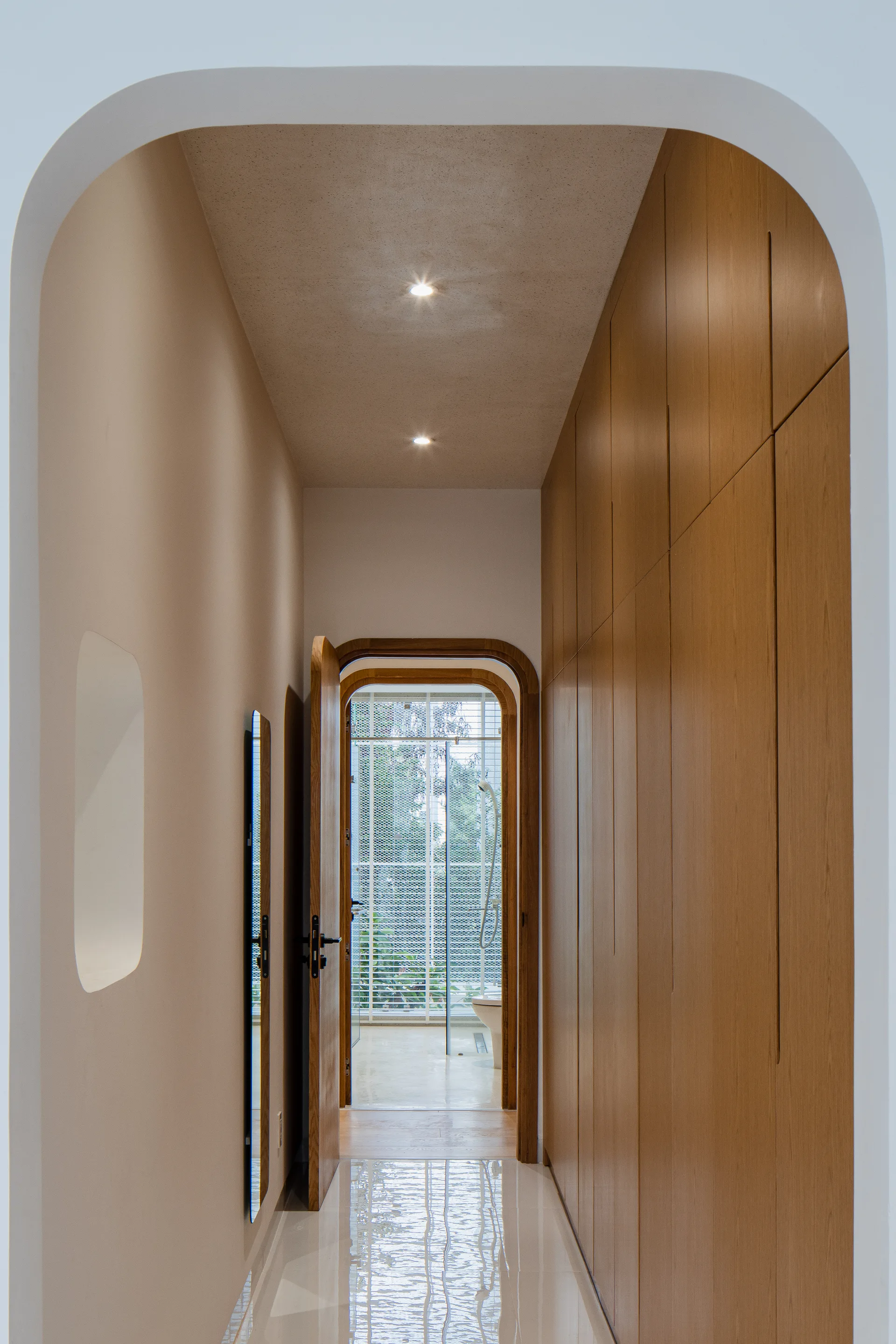 A minimalist hallway features built-in light wood cabinetry, a polished beige floor, a slim wall mirror, and rounded plaster archways.