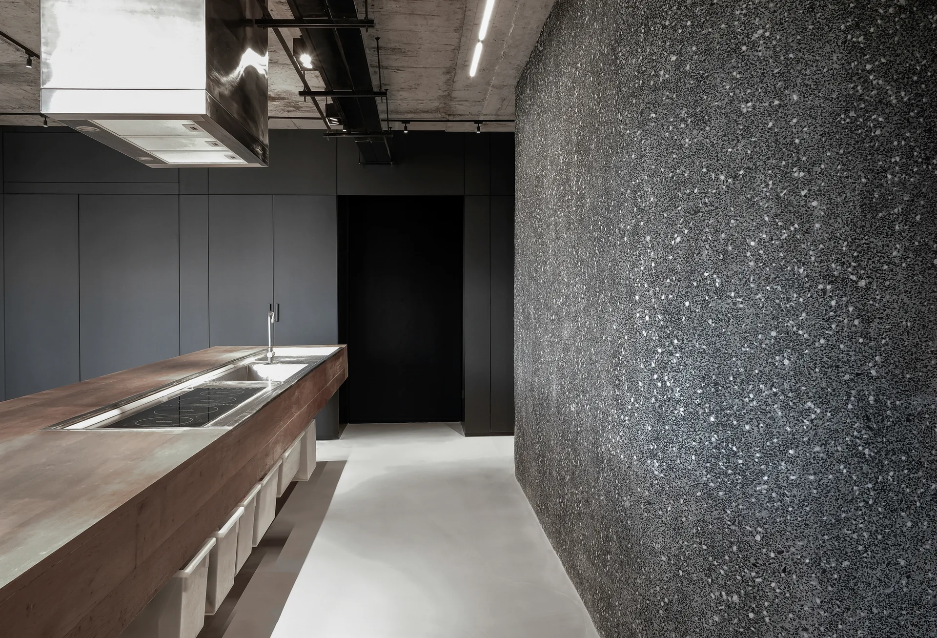 A minimalist industrial kitchen featuring a wooden island with an integrated sink, a large grey terrazzo wall, and a raw concrete ceiling.
