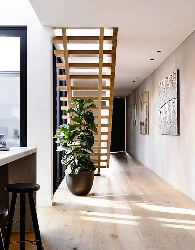 An open-tread wooden staircase stands in a bright hallway with light wood floors, a potted fiddle leaf fig plant, and contemporary wall art.