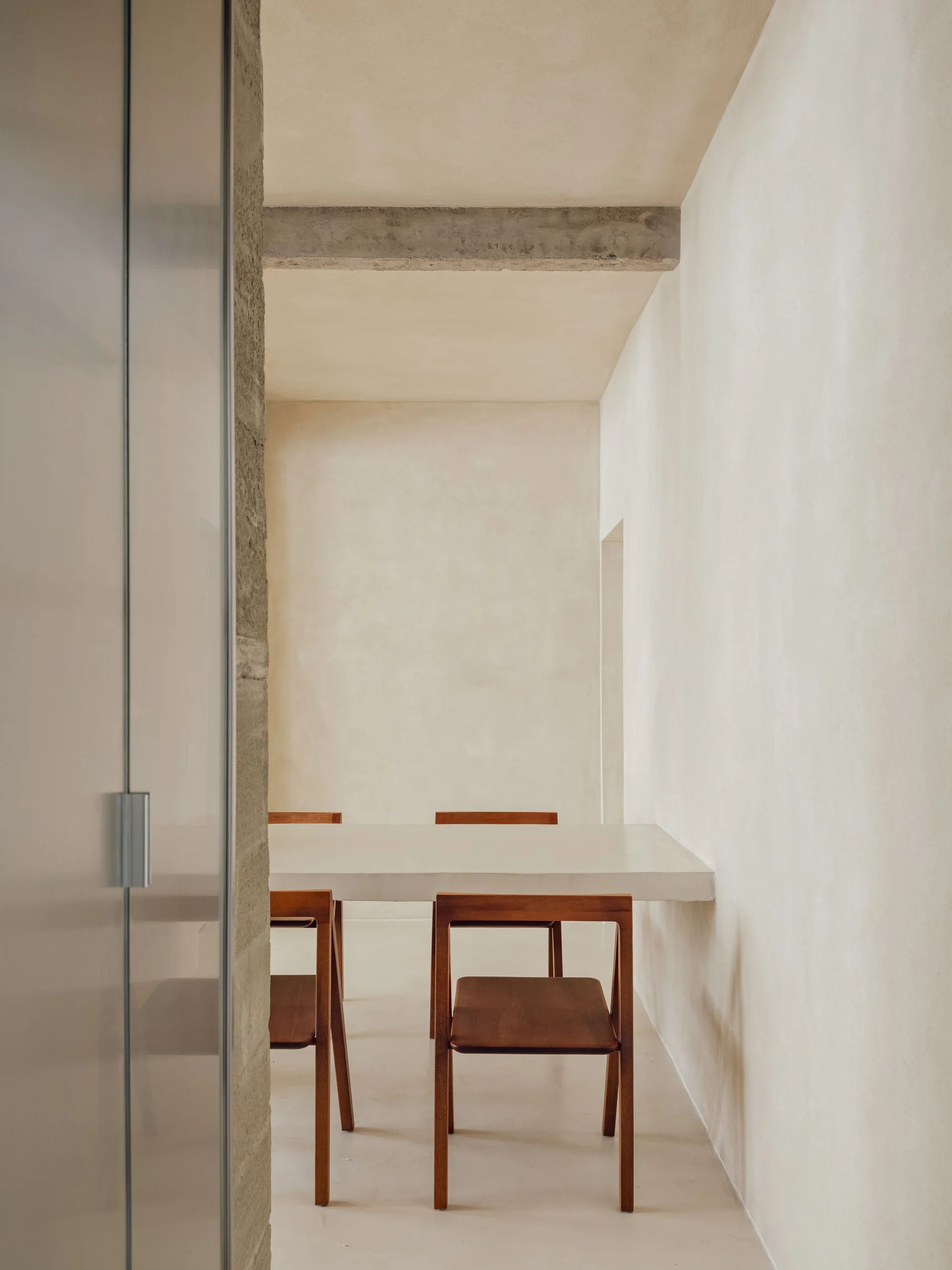 A minimalist dining area features wood chairs surrounding a white table beneath a concrete ceiling beam and warm beige plaster walls.