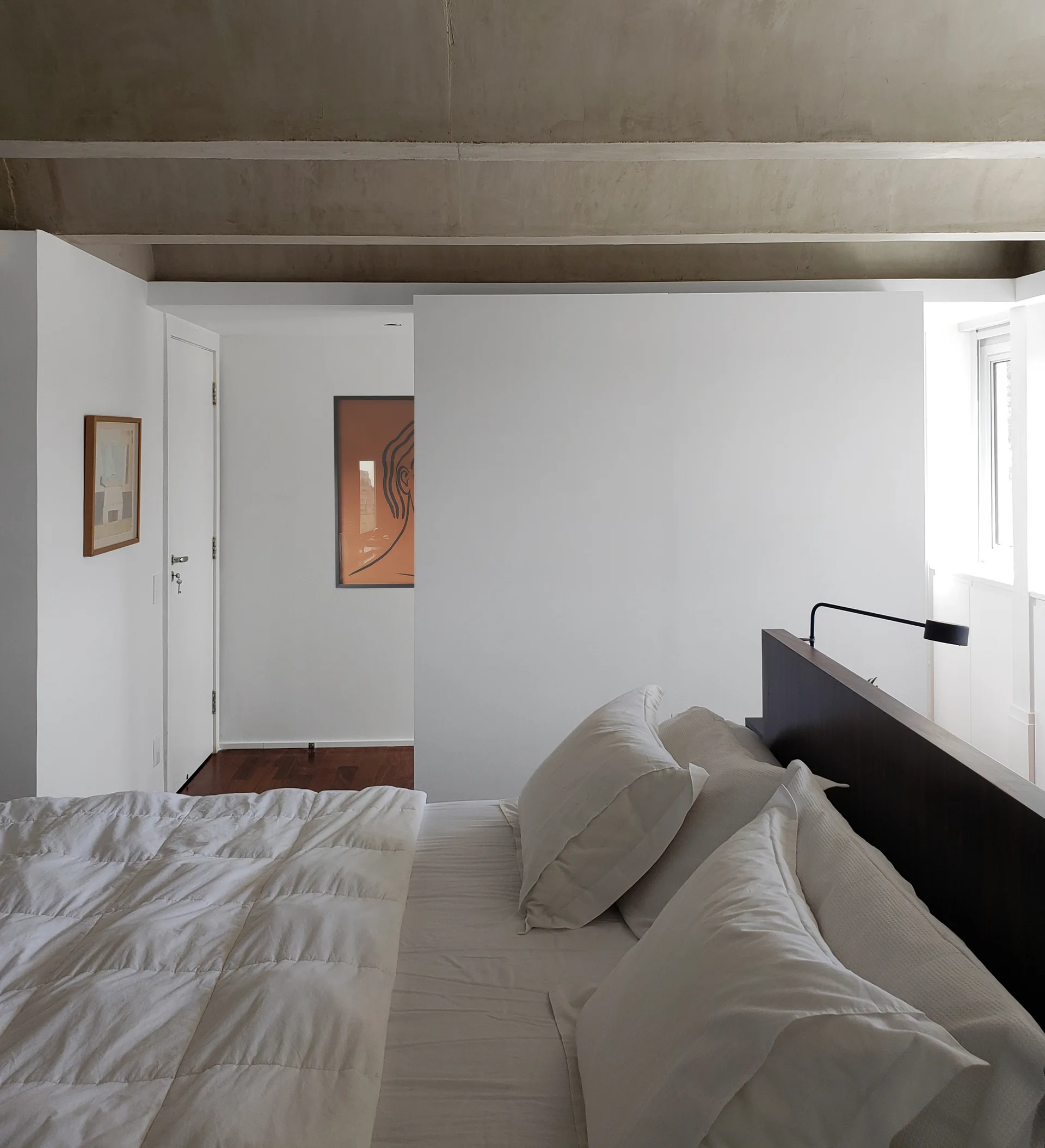 A minimalist bedroom featuring a bed with white linens beneath an exposed concrete beam ceiling and next to a white partition wall.