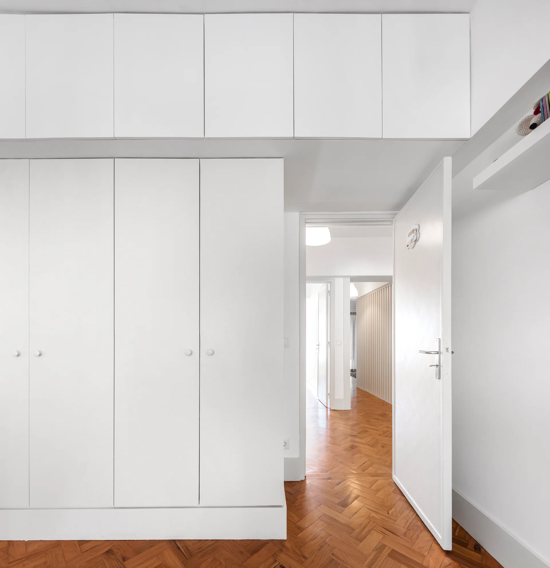 A bright hallway features floor-to-ceiling white built-in cabinets and a classic warm wood herringbone floor with an open doorway leading to another room.
