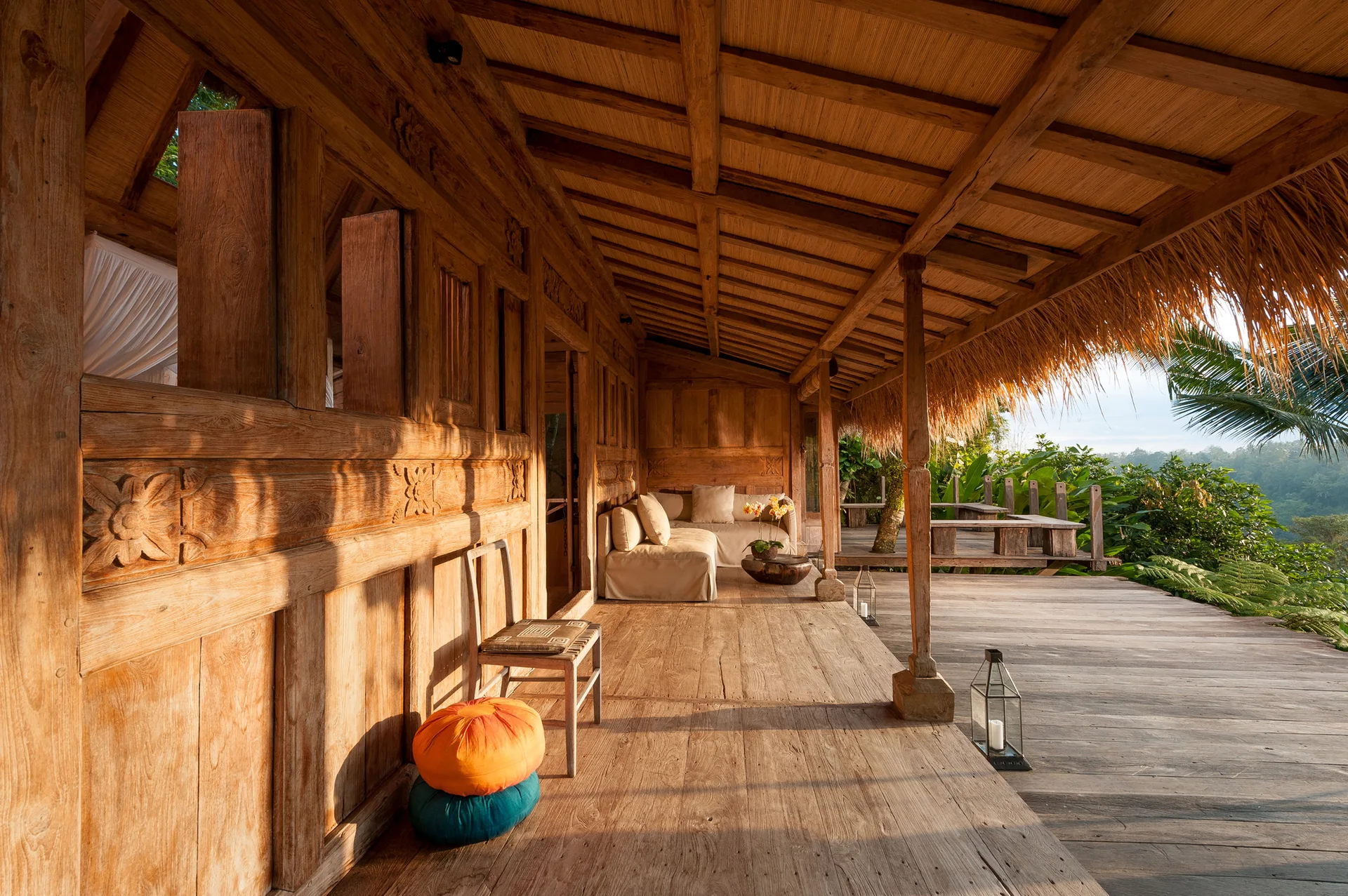 A wide wooden porch features hand-carved walls, a thatched roof, a neutral-toned sofa, and colorful floor cushions on the weathered deck.