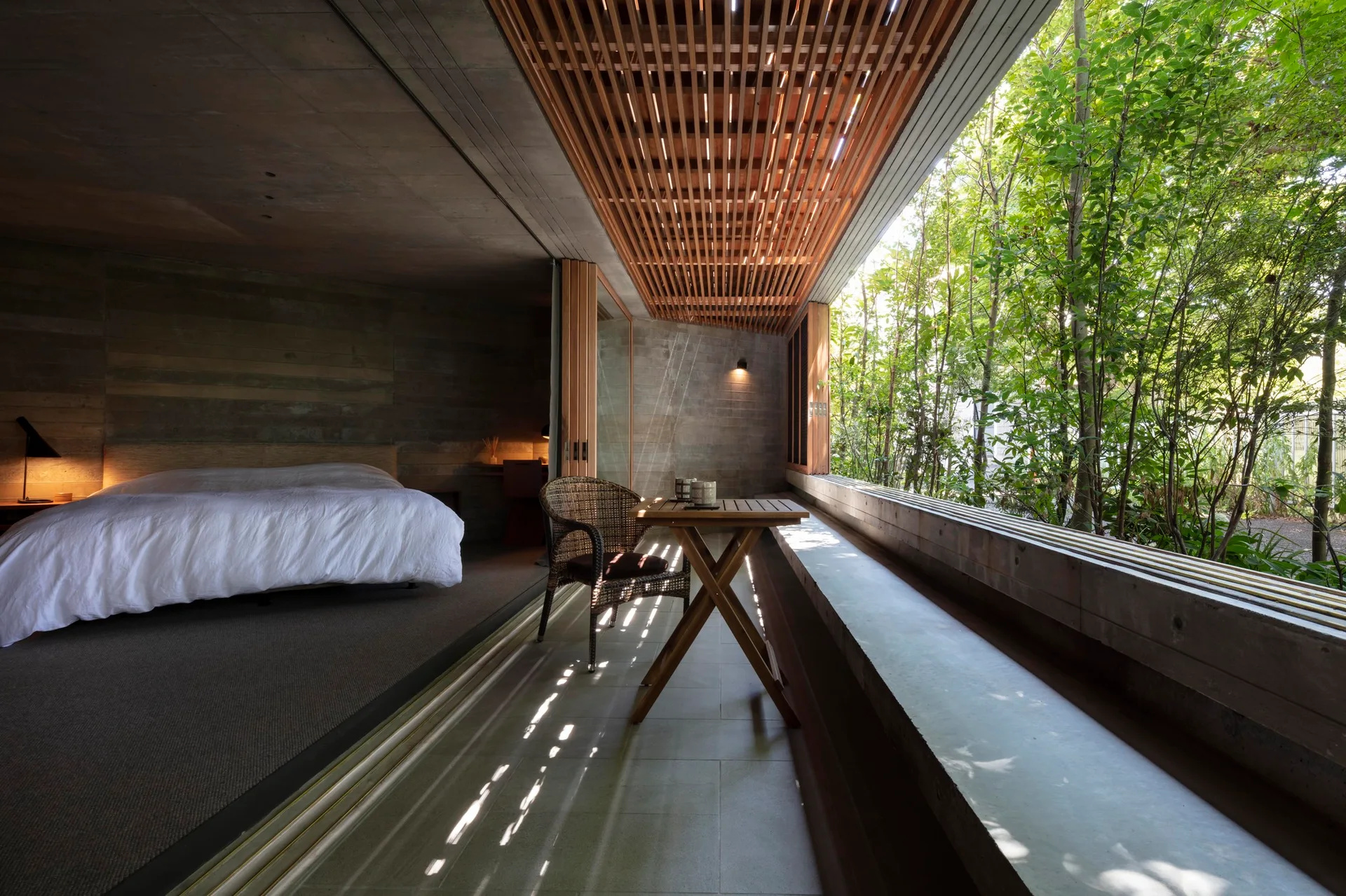 A modern bedroom with concrete walls and white bedding opens to a semi-outdoor tiled patio with a wood slatted ceiling and folding glass doors.