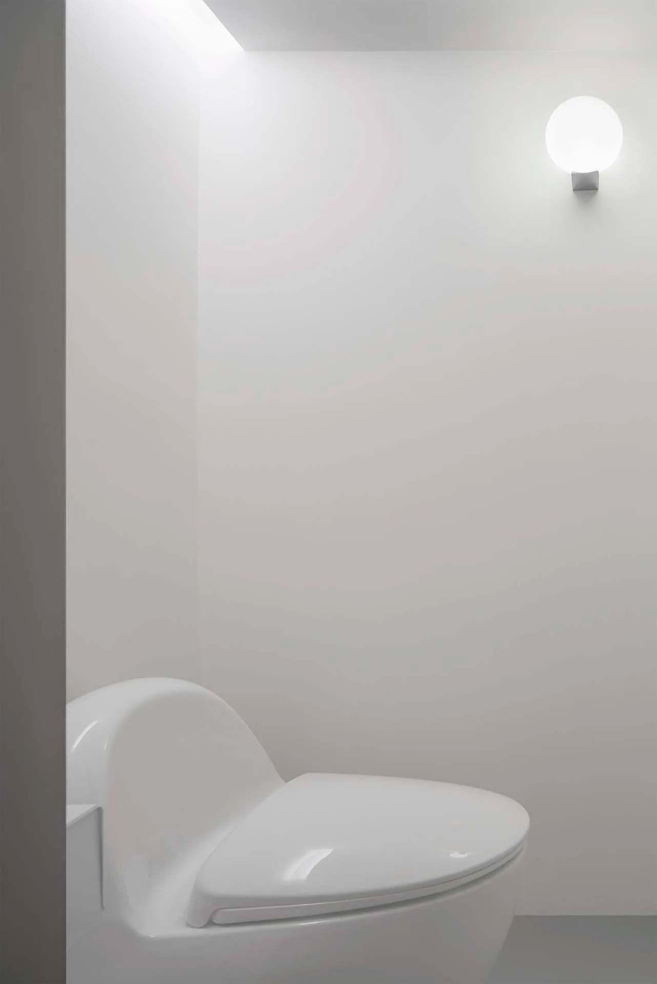 A stark white powder room features a curved white toilet next to a smooth wall with an illuminated circular globe sconce.