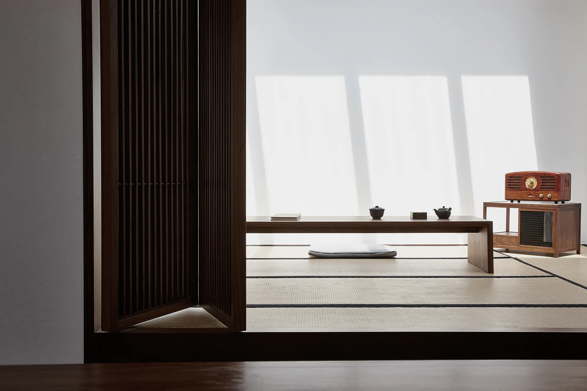 A minimalist room featuring tatami mat flooring, a long low wooden table with tea items, and a dark wood slatted screen door.