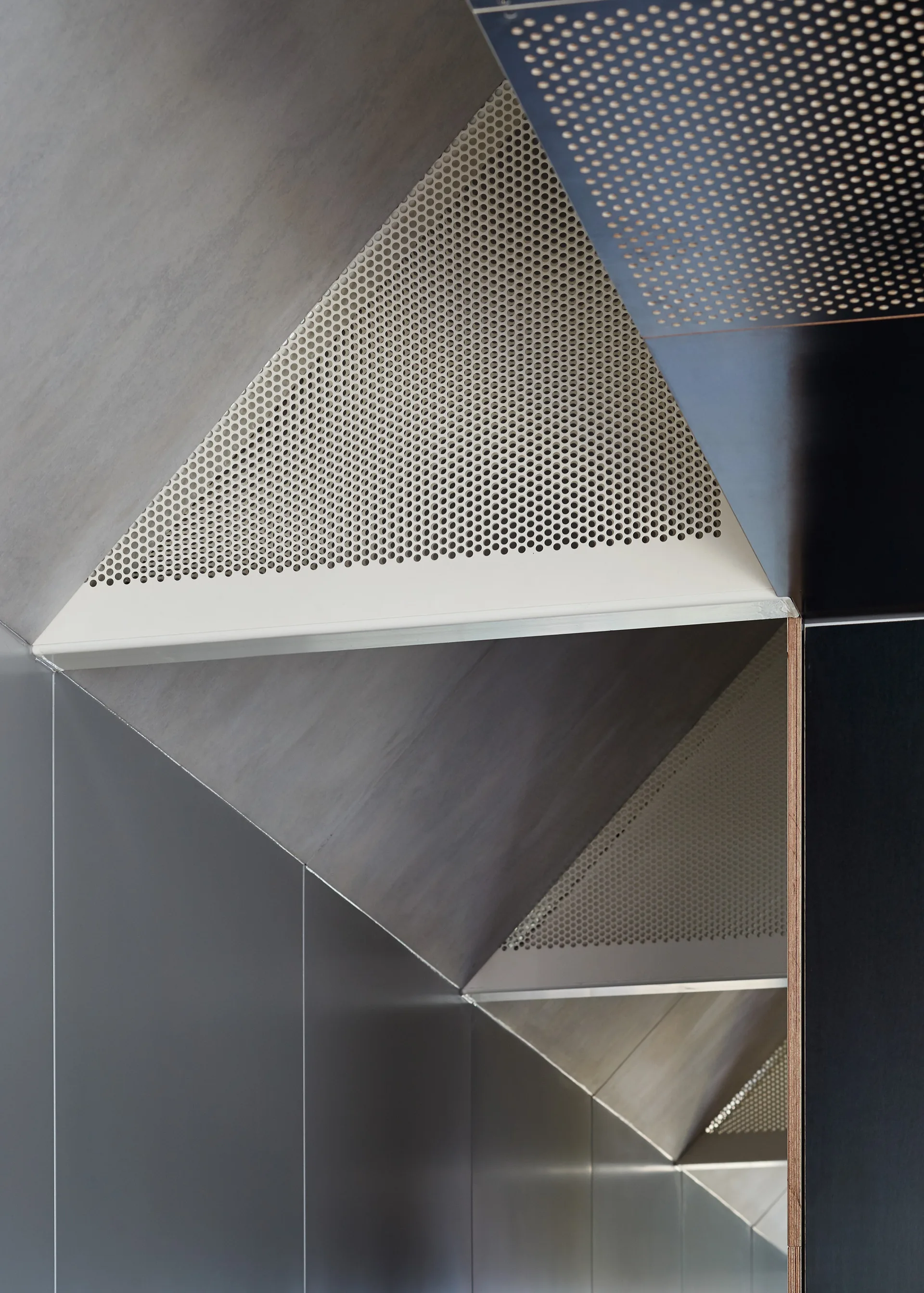A close-up of geometric architectural paneling with sharp angles, featuring satin grey metal and white perforated triangular panels.