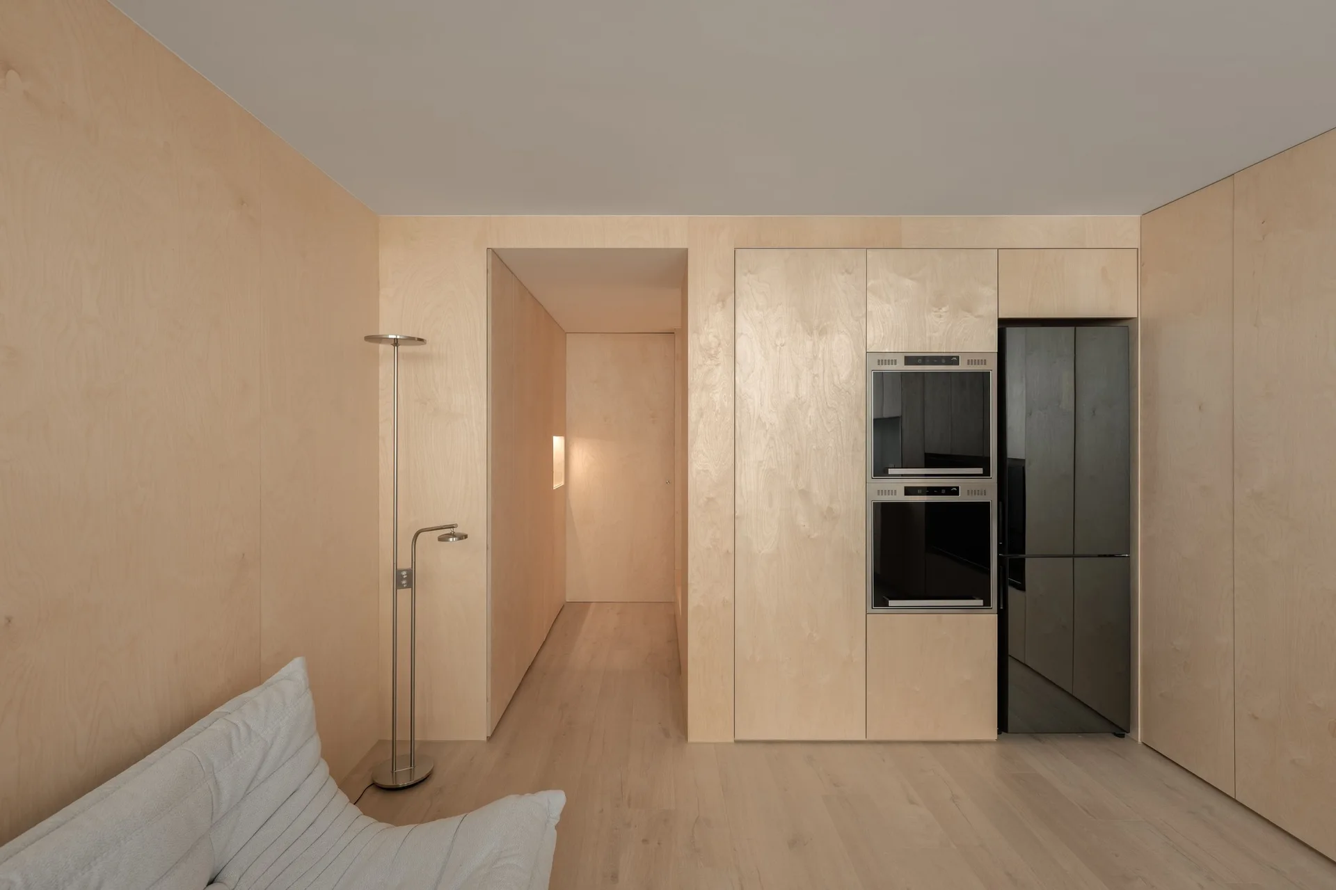 Minimalist interior featuring extensive light wood paneling on the walls and kitchen cabinets, with a black built-in oven, refrigerator, and silver floor lamp.