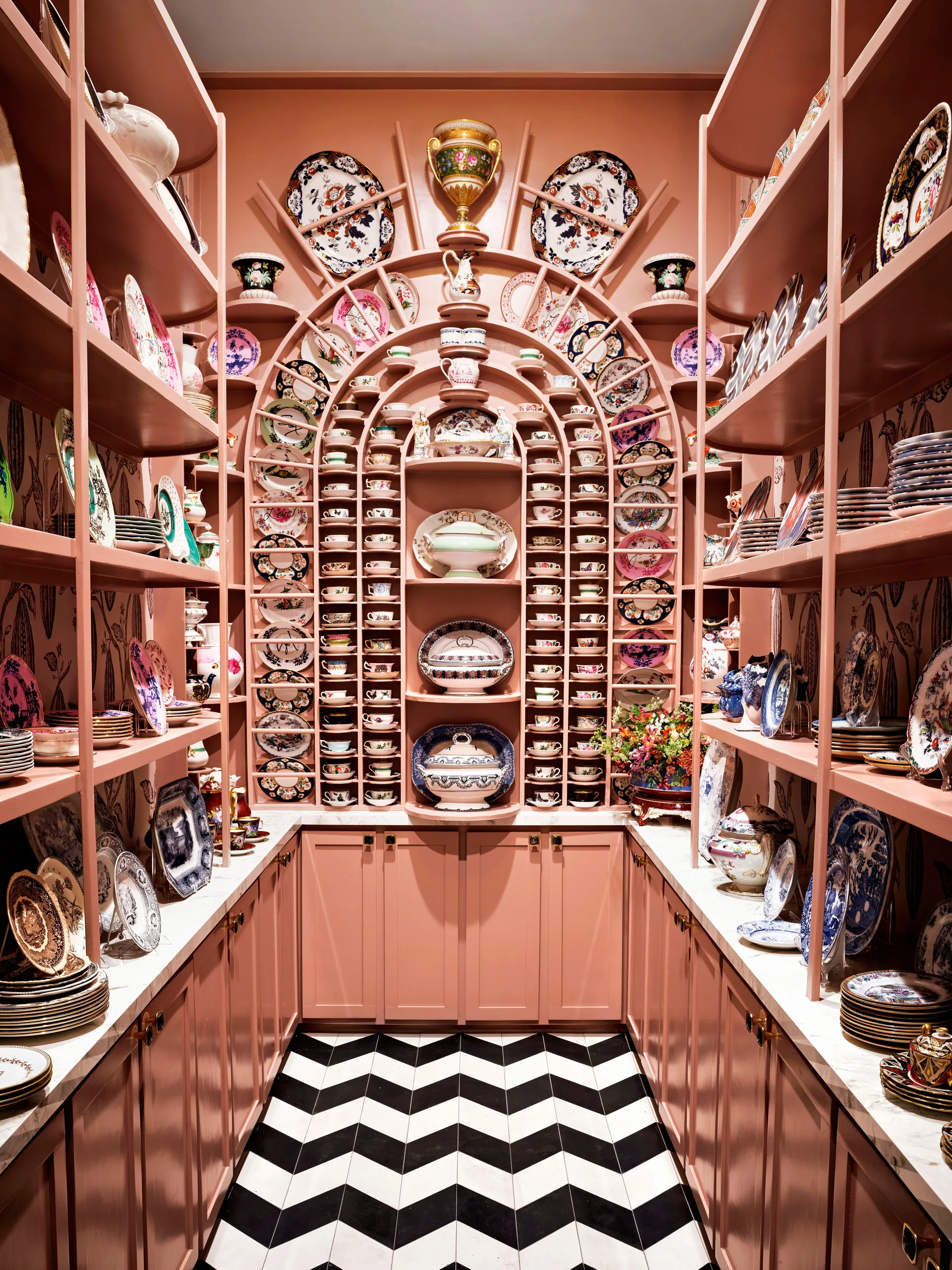 Symmetrical pink butler's pantry featuring floor-to-ceiling built-in shelving packed with decorative china over a black and white chevron floor.