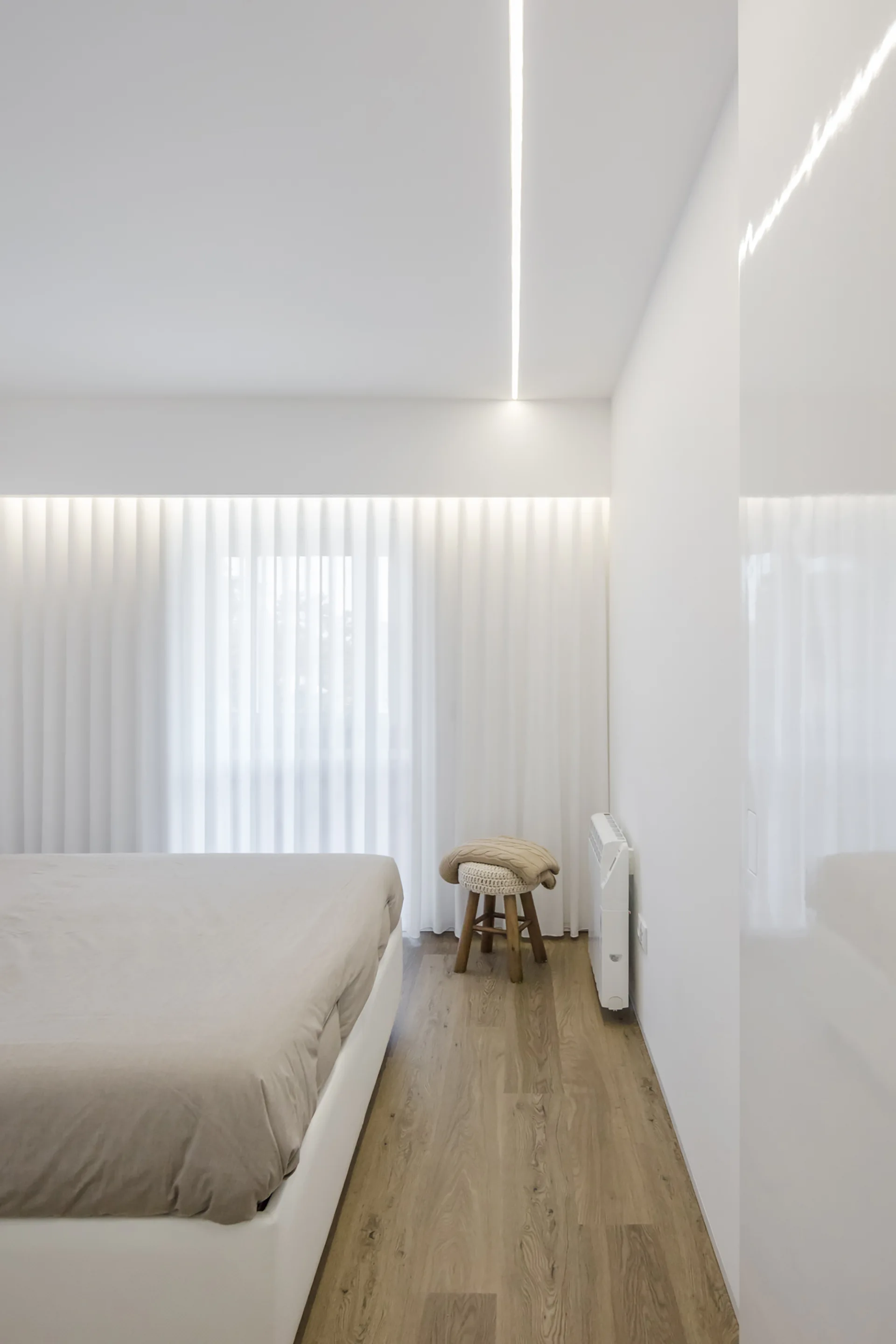 A minimalist bedroom features a bed with beige sheets, white walls, wood floors, sheer curtains, and a glowing linear light strip on the ceiling.