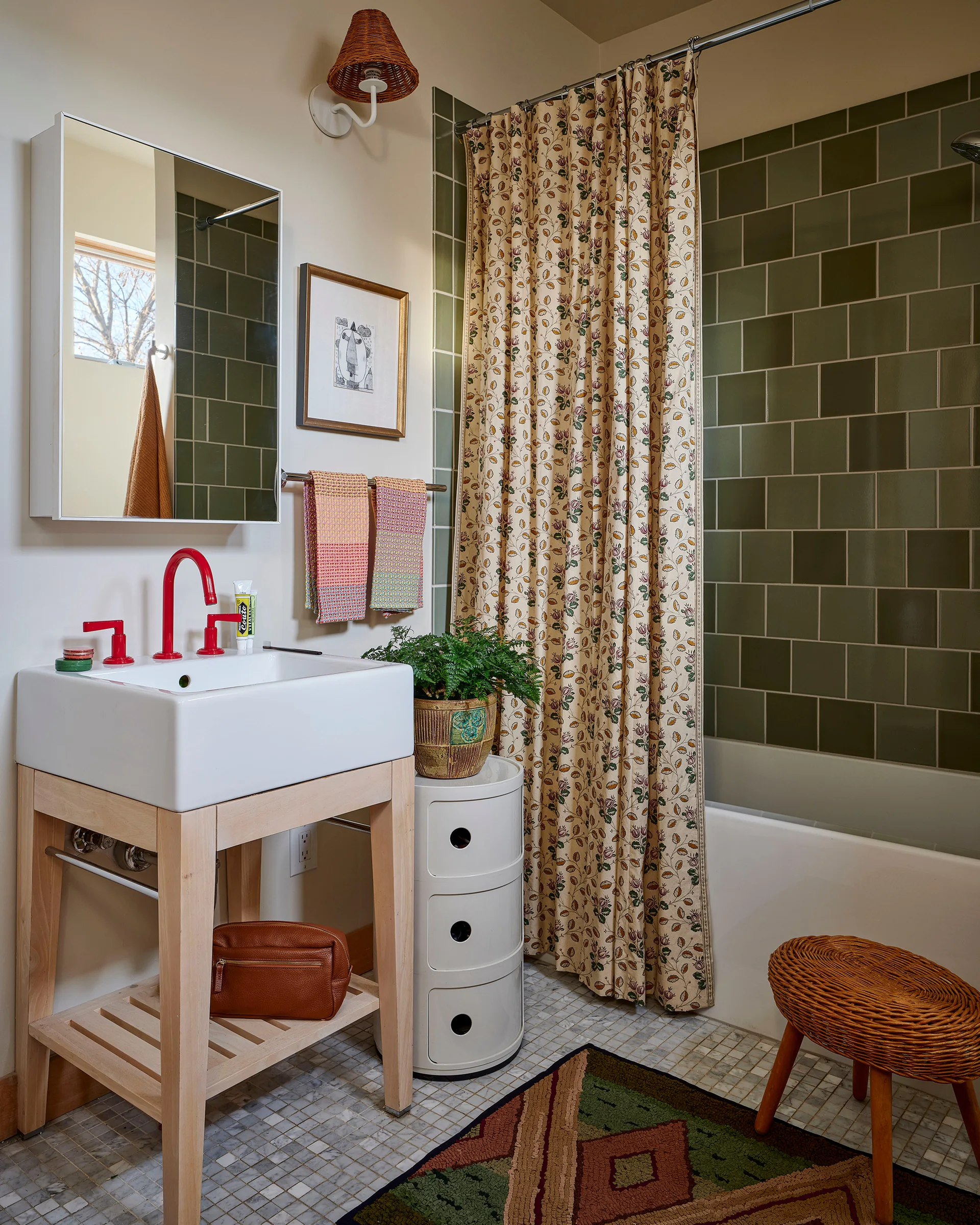 An eclectic bathroom features a light wood vanity with a white sink and red faucet, a floral shower curtain, and sage green wall tiles.