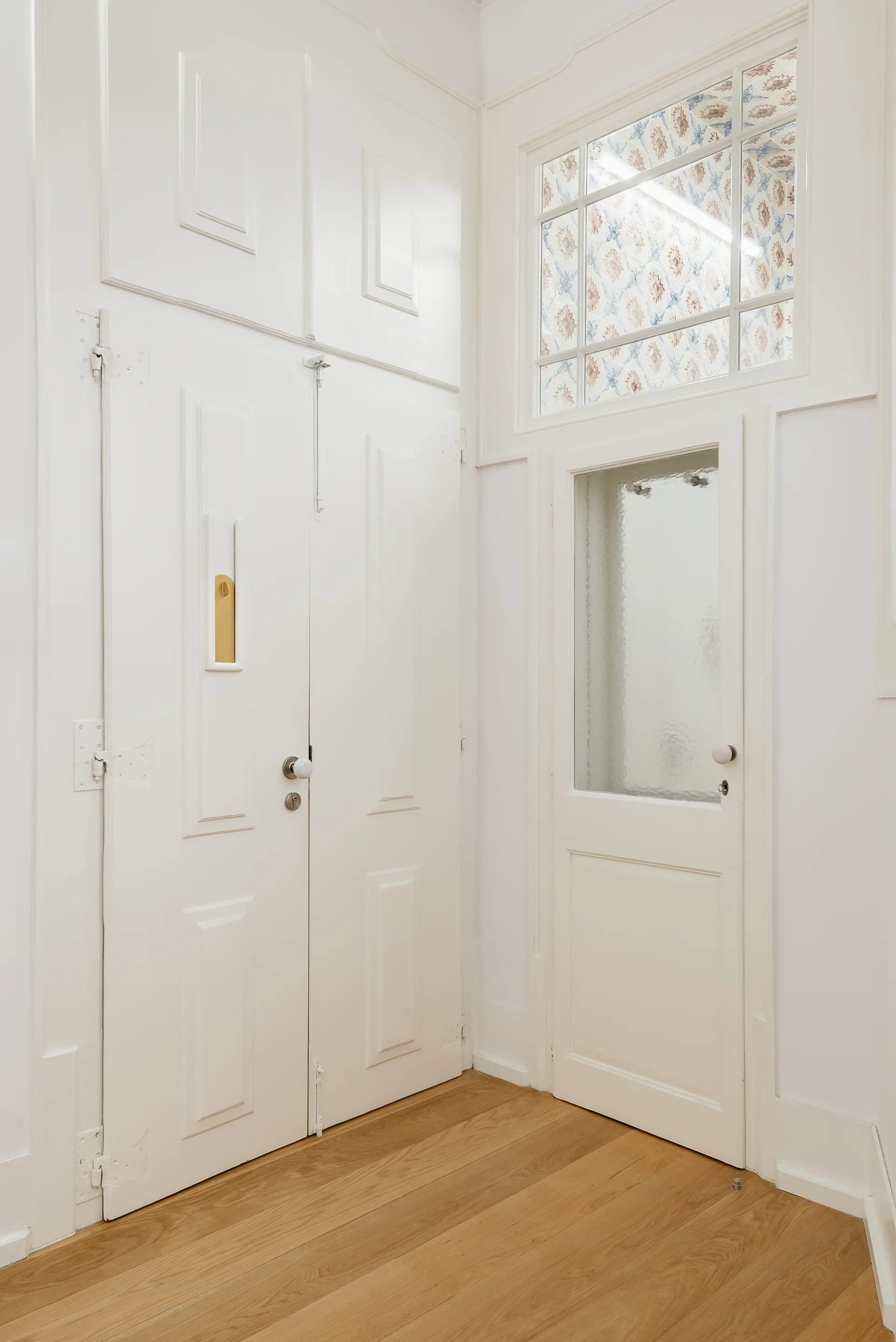 White painted paneled doors and a glass-paneled door with a patterned transom window stand on a light hardwood floor.