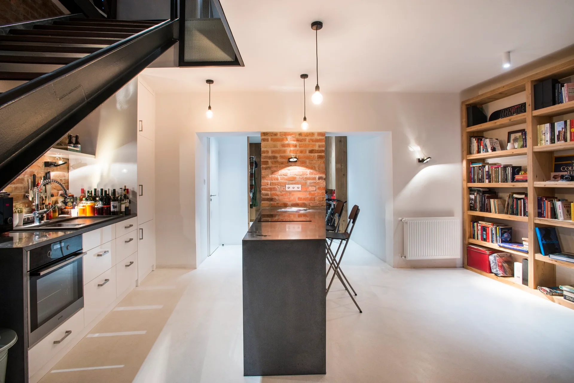 A modern kitchen featuring a dark central island, white cabinets under a metal staircase, and a wooden bookshelf set against a white wall.