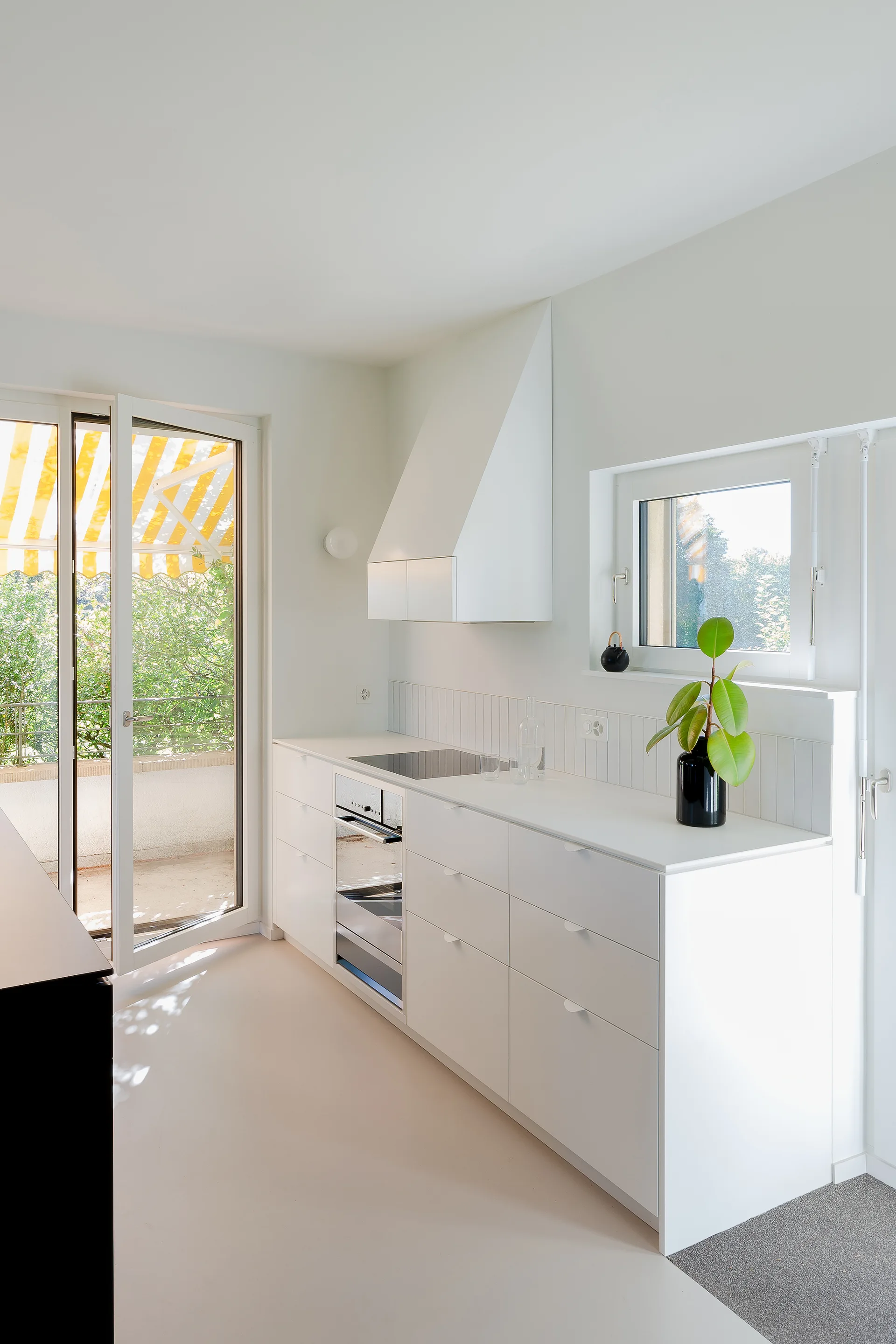 A minimalist white kitchen features flat-front base cabinets, a vertical tile backsplash, a white angled range hood, and a glass door opening to a balcony.