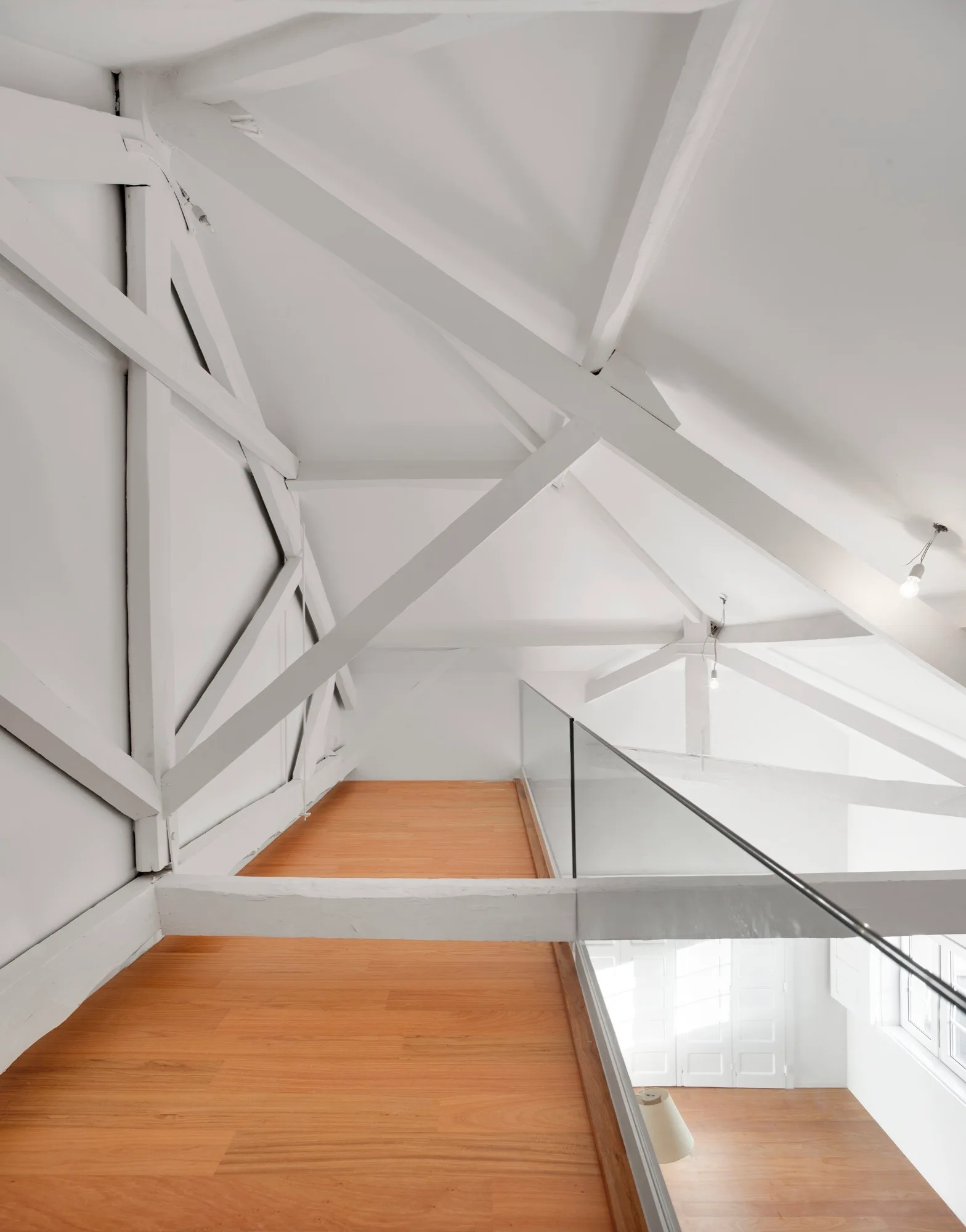 A bright attic loft with warm wood flooring, a transparent glass railing, and structural wooden roof beams painted entirely in matte white.