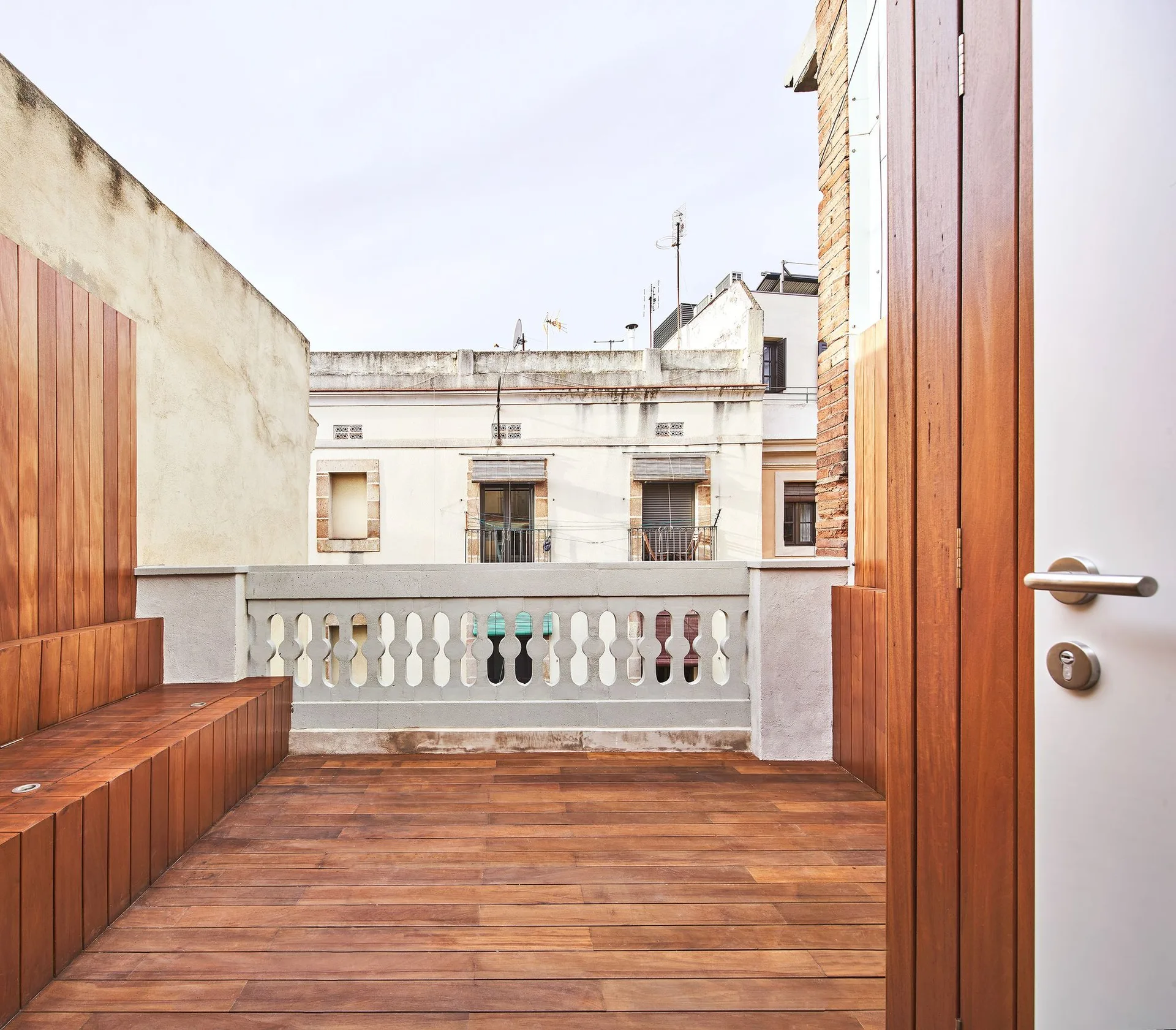An urban terrace features rich warm wood decking, a built-in tiered wooden bench, a stone balustrade, and a partially open wooden door.