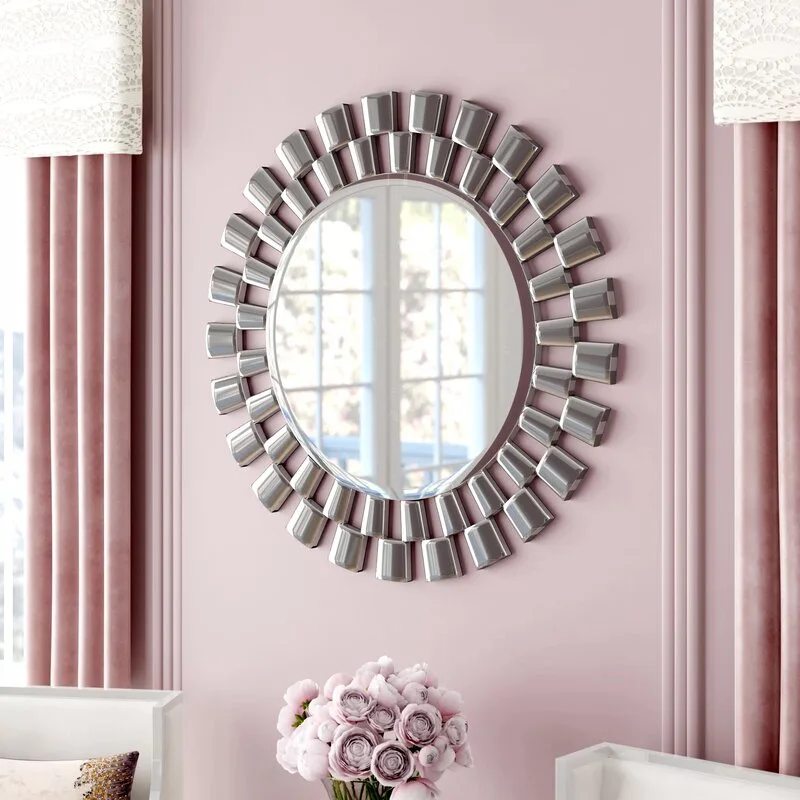 A decorative round mirror with a metallic sunburst frame hangs on a soft pink paneled wall between two pink velvet curtains.