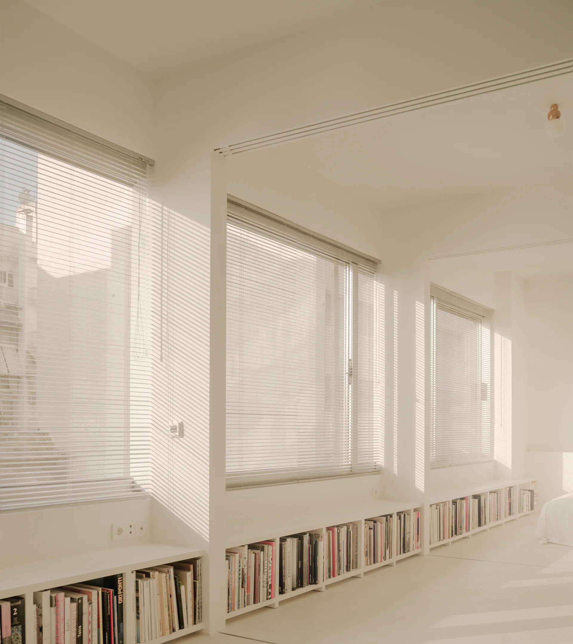 A low, continuous white bookshelf filled with books sits beneath a row of windows with Venetian blinds in a minimalist white room.