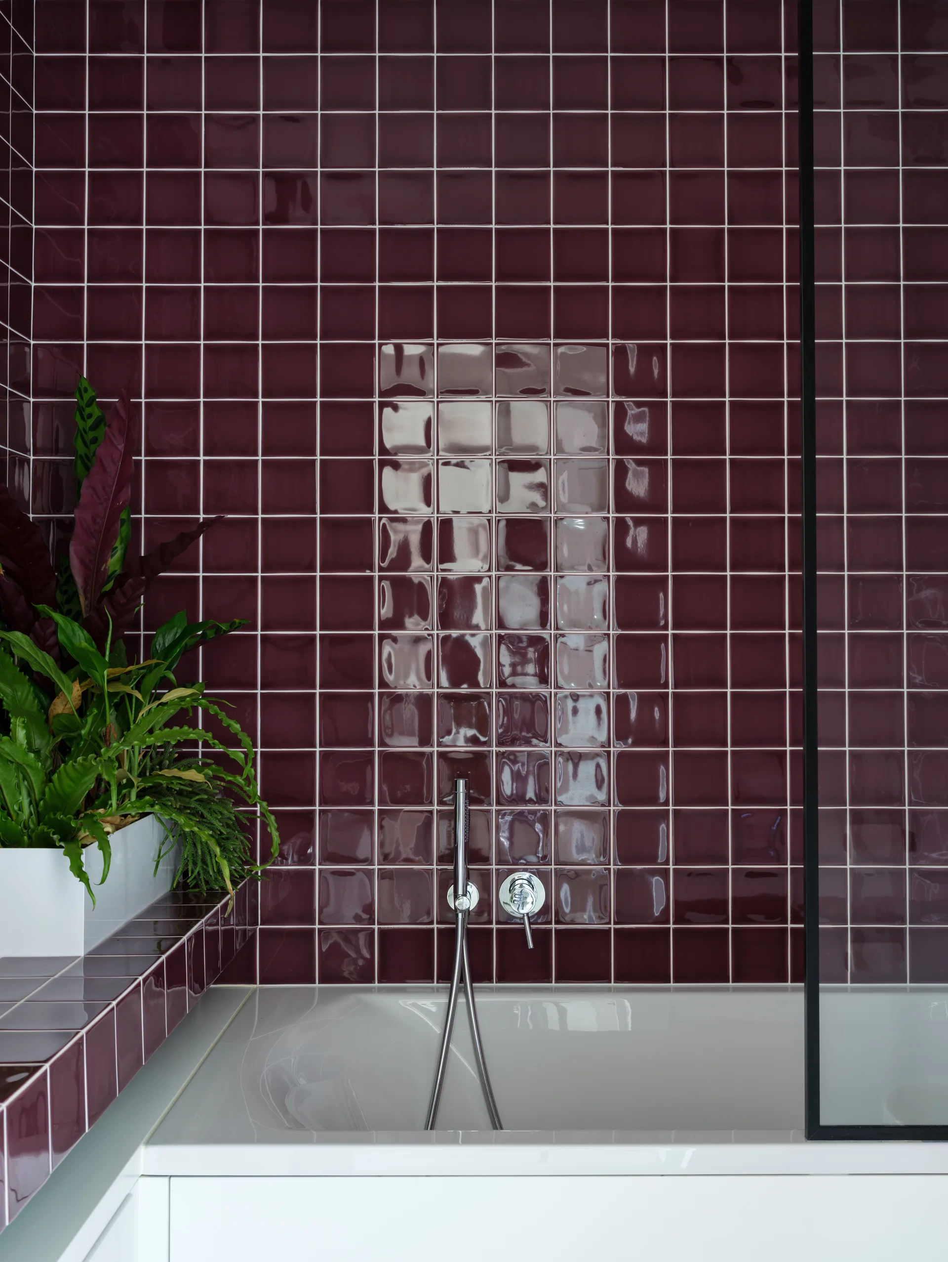 A white bathtub sits against a wall of glossy burgundy square tiles with a chrome faucet and a green potted plant on the ledge.