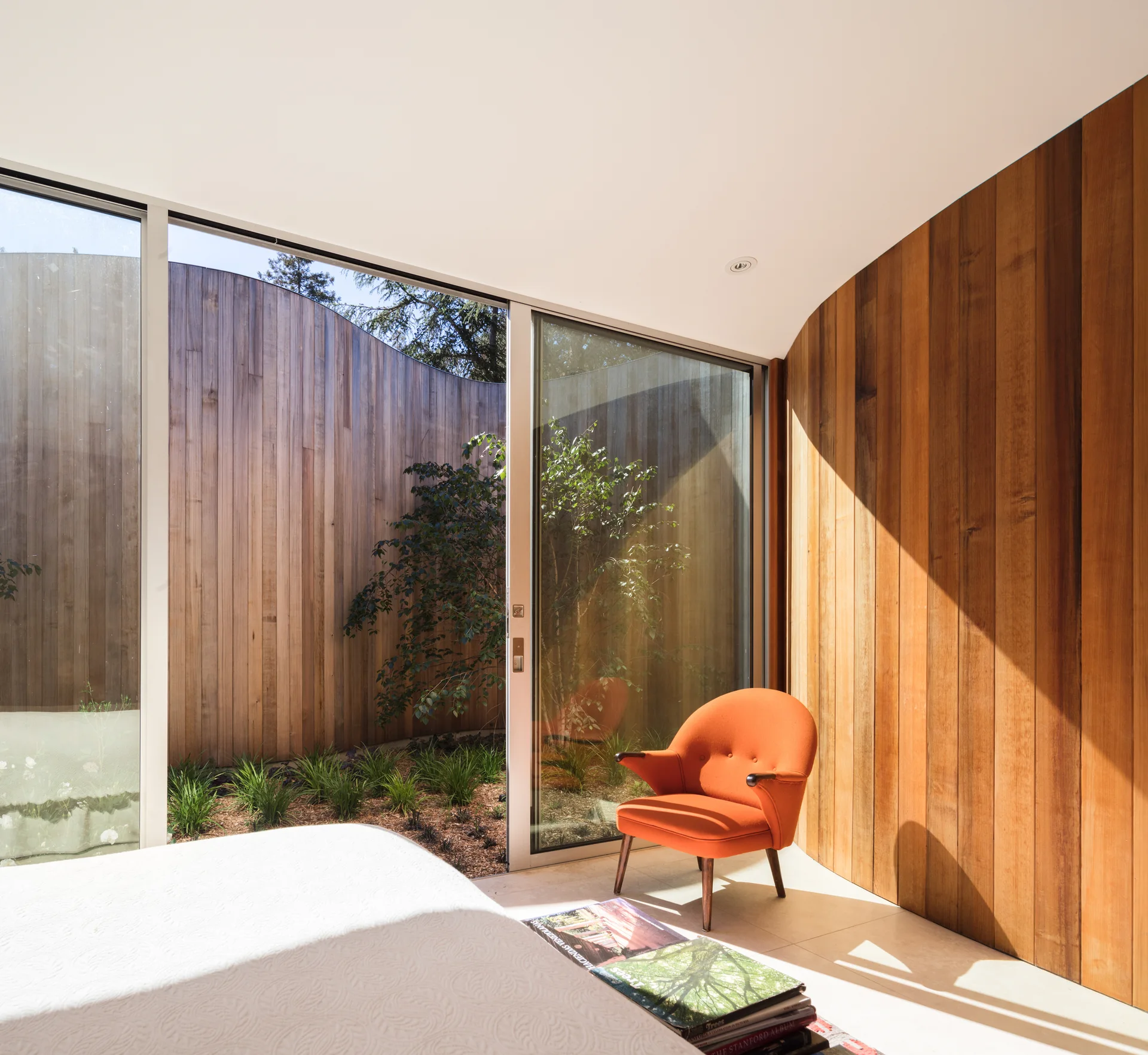 A modern bedroom corner features a curved vertical wood plank wall, an orange lounge chair, and glass sliding doors opening to a wood-walled courtyard.