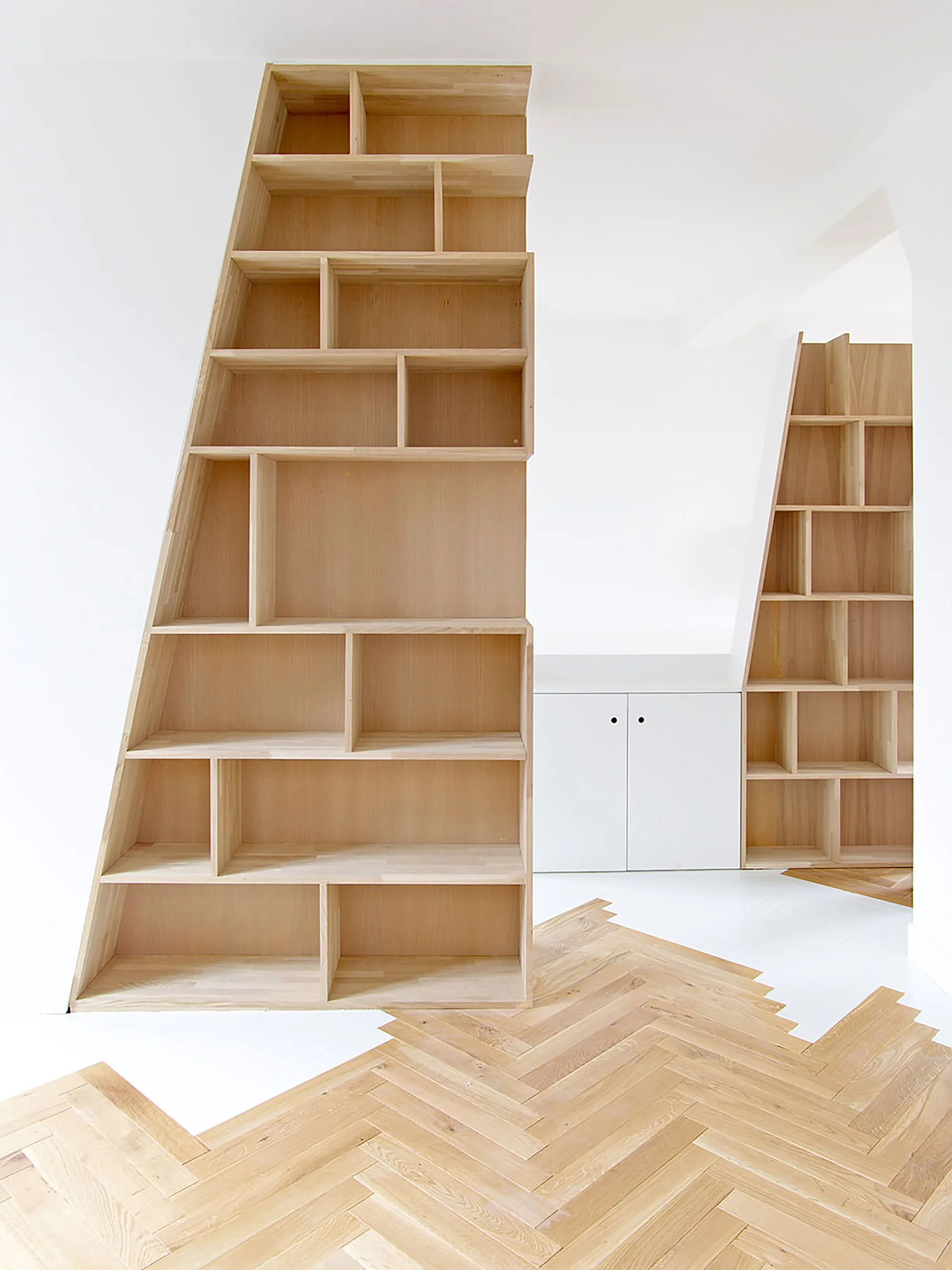 Two minimalist light wood bookshelves with slanted sides stand on a unique herringbone wood floor that transitions jaggedly into white flooring.