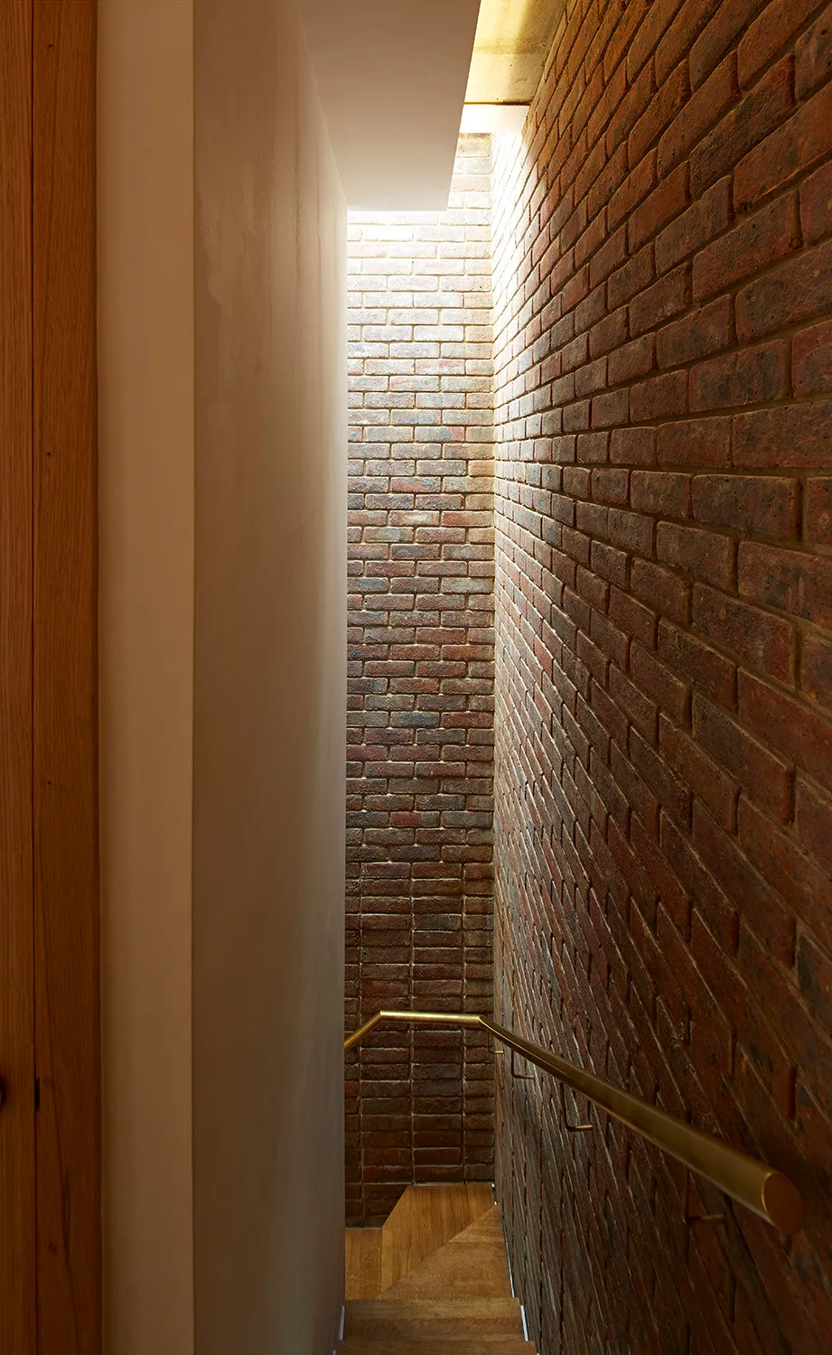 A narrow staircase with wooden treads features an exposed red brick wall, a gold metal handrail, and bright natural light from an overhead skylight.