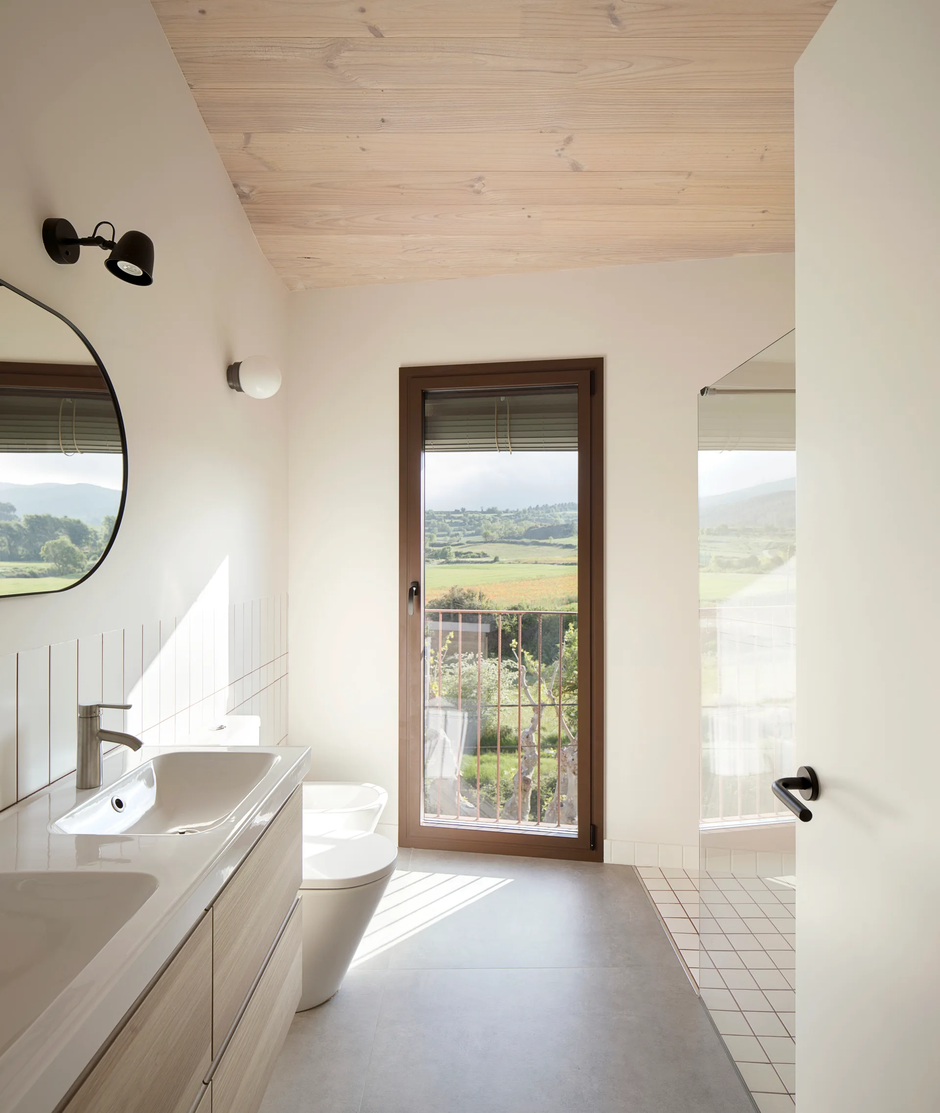 A minimalist bathroom featuring a light wood vanity, a wood-plank ceiling, a circular mirror, and a tall glass door showcasing a rural landscape.