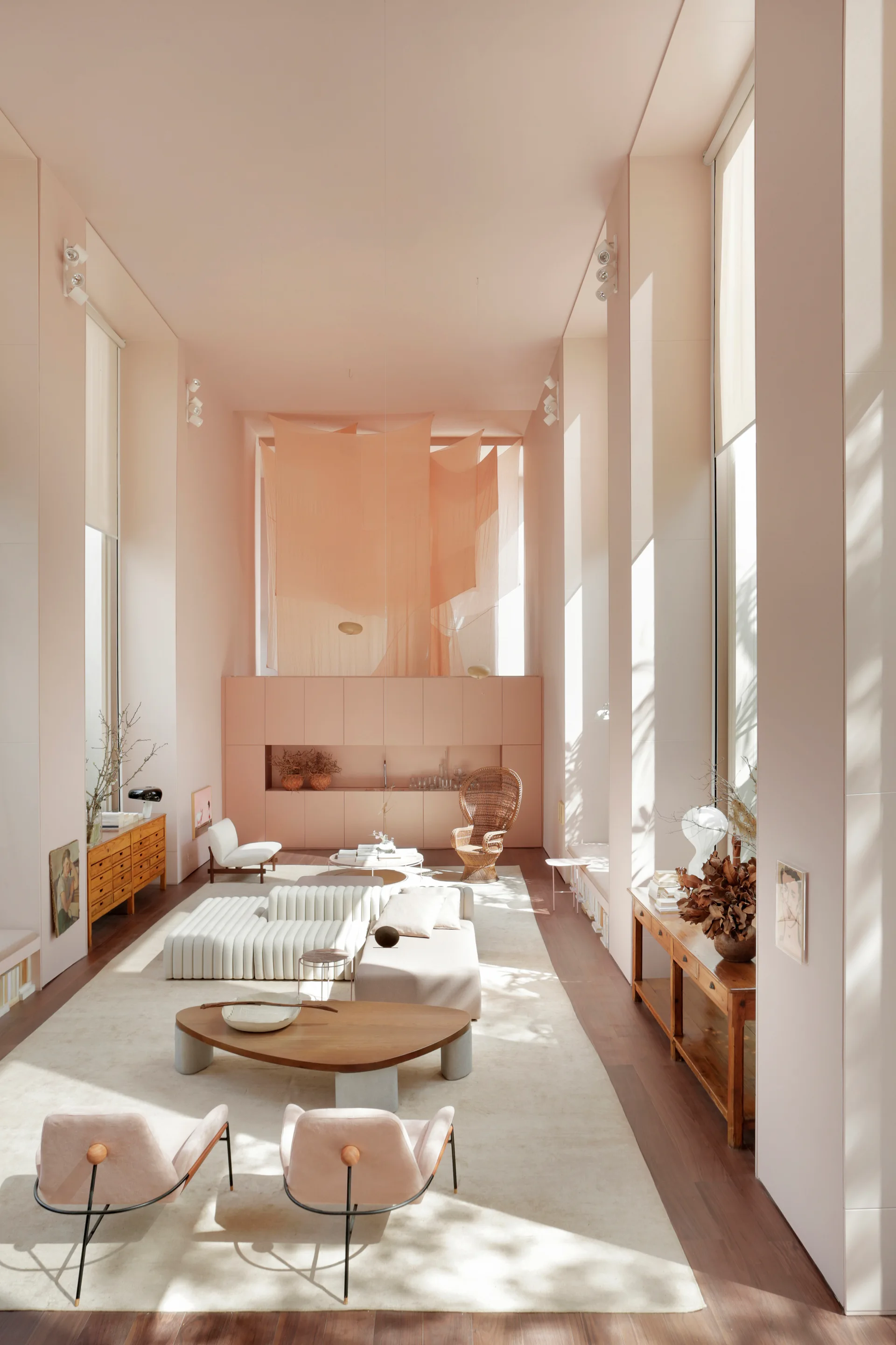 A high-ceilinged living room with peach walls features a low-profile white sectional sofa, wooden coffee tables, and draped fabric overhead.