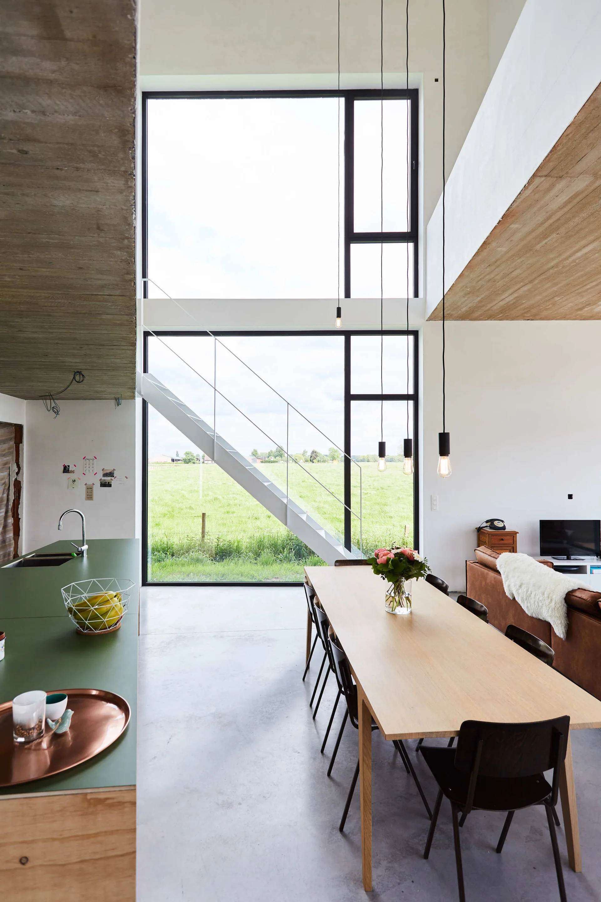 A light wood dining table with black chairs sits in an open-plan room with double-height windows, a concrete ceiling, and a white open staircase.
