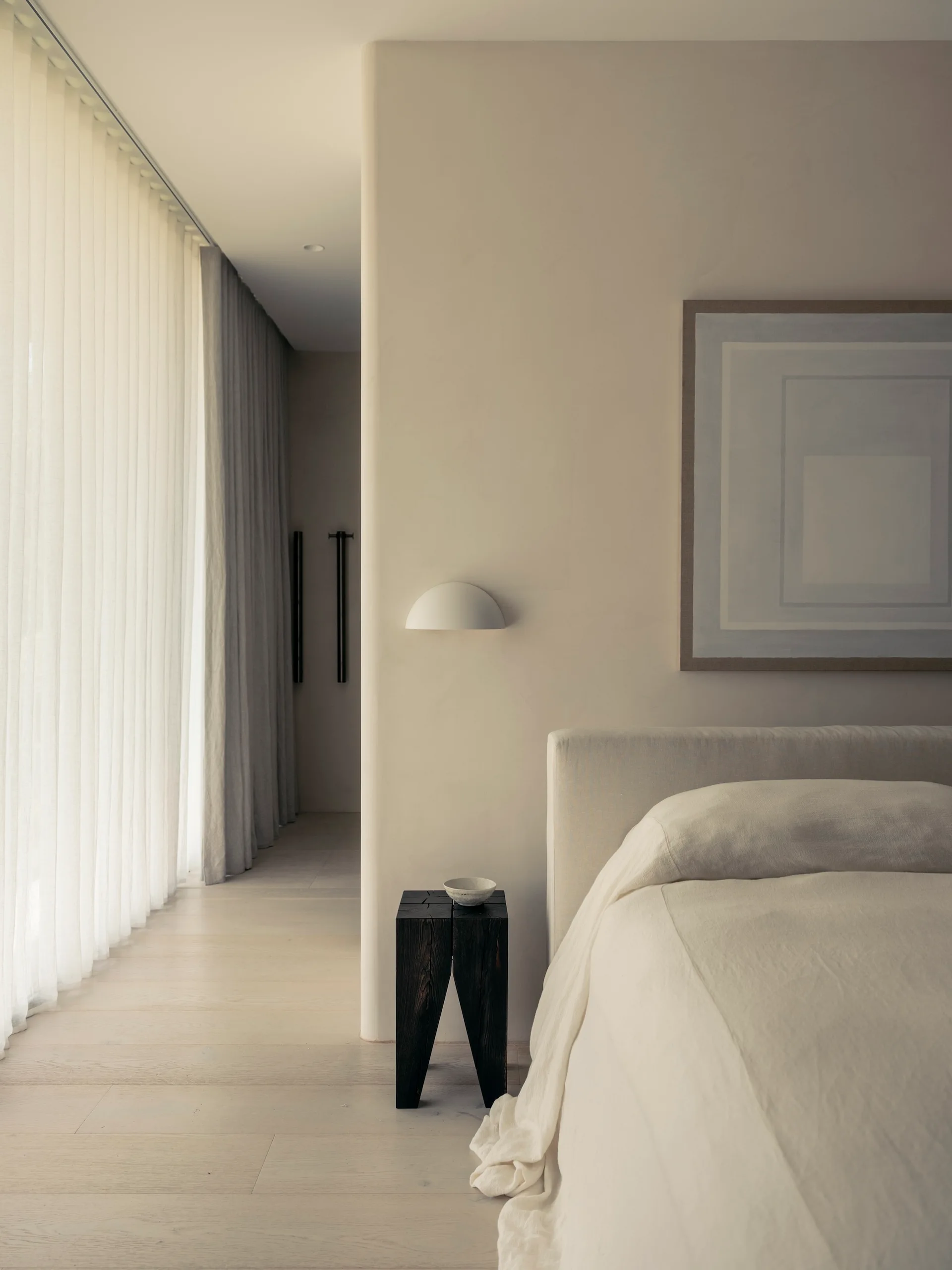 A minimalist beige bedroom features a linen-covered bed, a dark wood block side table, a white dome sconce on a curved plaster wall, and sheer curtains.