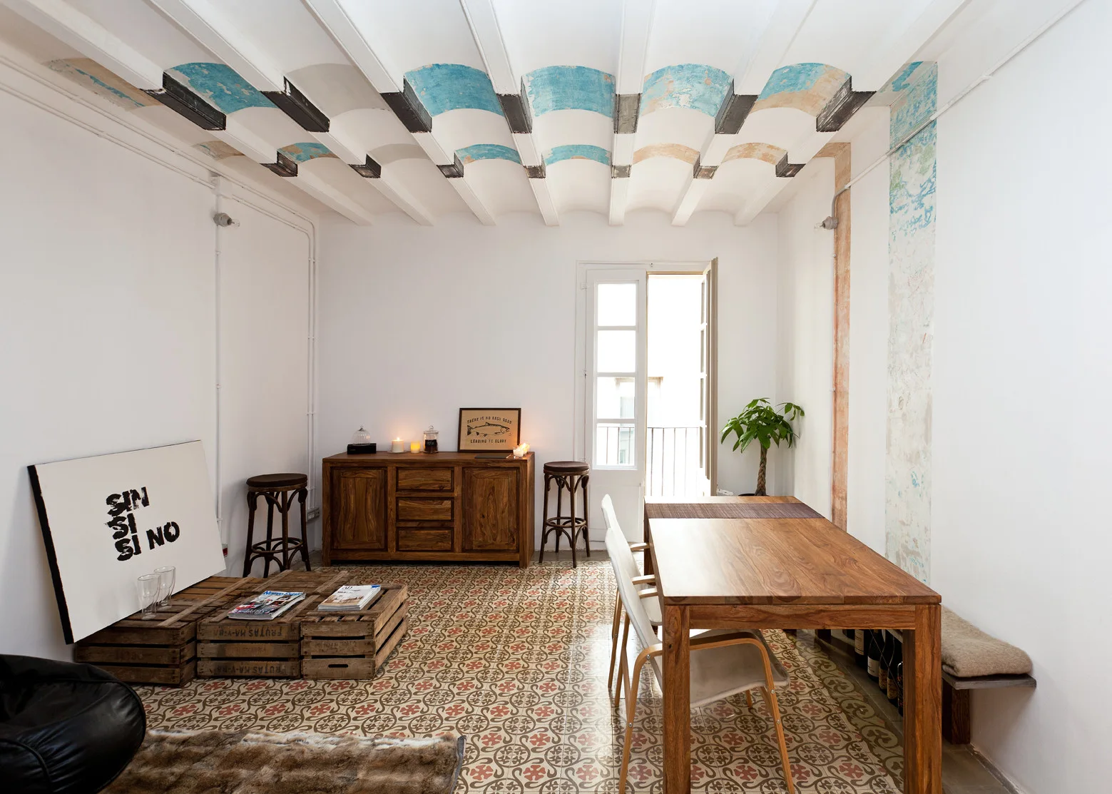 A rustic dining room features a wooden table, a matching sideboard, patterned ceramic floor tiles, and a vaulted ceiling with blue arched details.