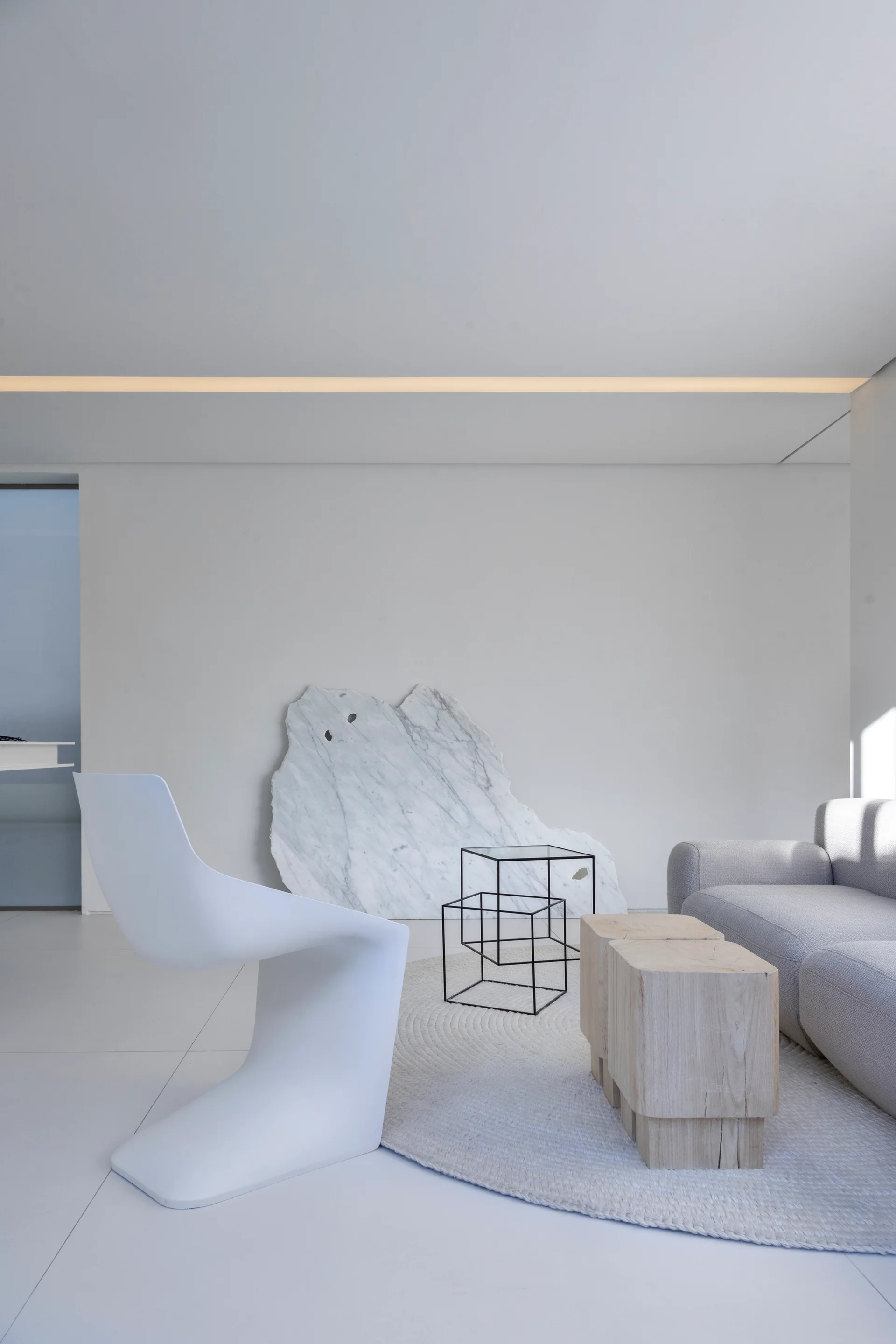 A minimalist living room features a white cantilever chair, a light gray sofa, block wood coffee tables, and a large raw marble slab leaning against the wall.