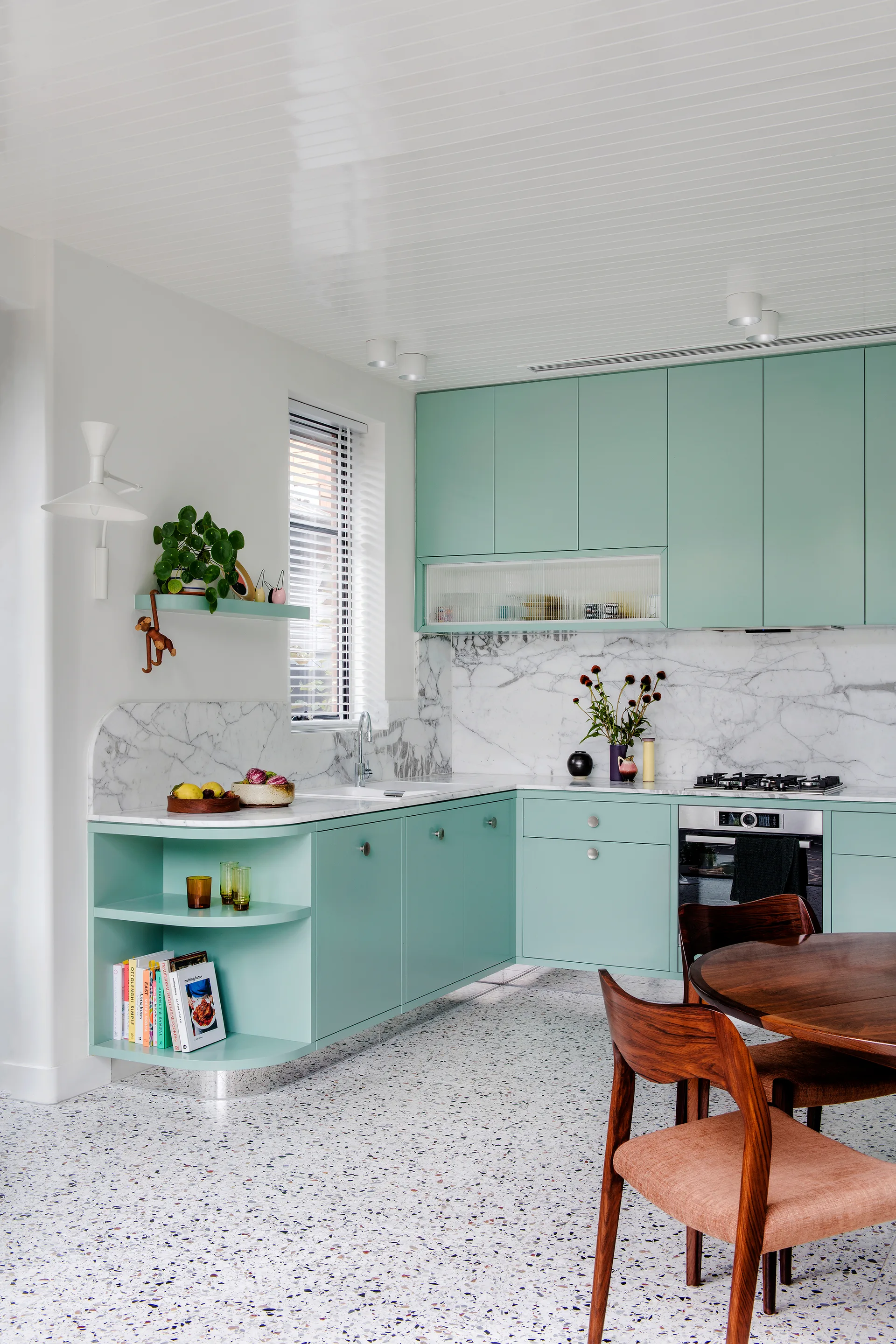 A modern kitchen features mint green cabinetry, a white veined marble backsplash, polished terrazzo flooring, and a dark wood dining table.