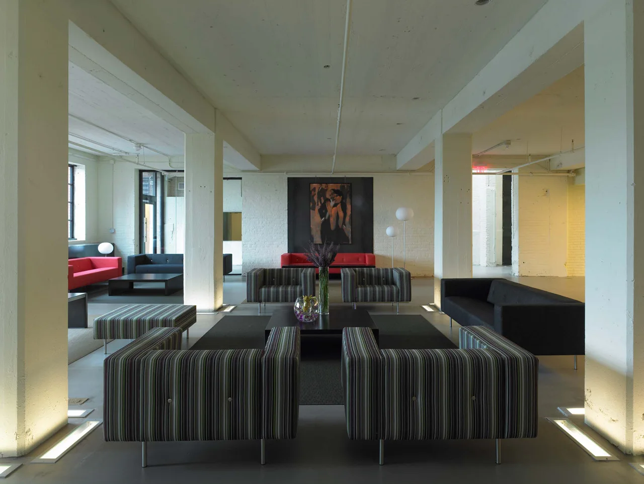 An industrial loft lounge features striped armchairs, dark sofas, and white concrete pillars illuminated by recessed floor uplighting.