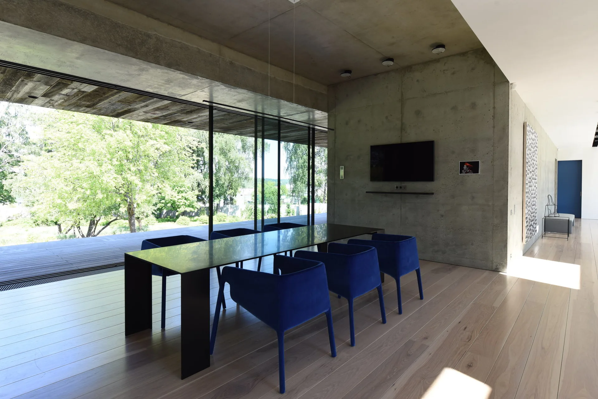 A minimalist dining area with a long dark table, vibrant blue chairs, an exposed concrete wall, and large sliding glass doors.