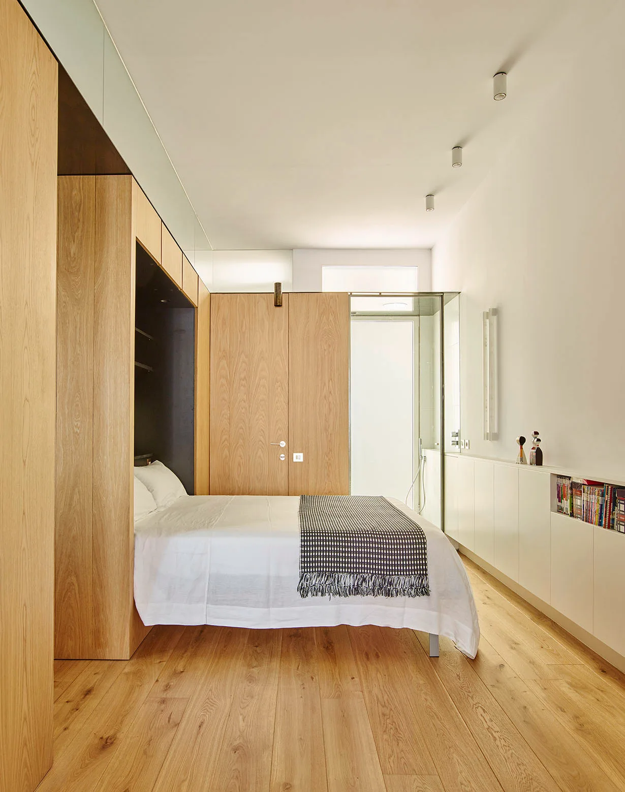 A minimalist bedroom featuring a bed recessed into light oak wood built-in cabinetry next to a low white credenza on matching wood floors.