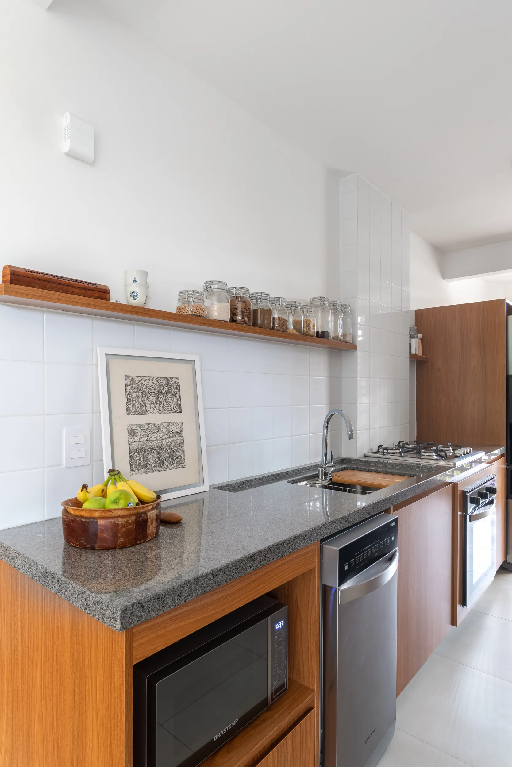 A modern kitchen counter with a gray granite countertop, wood-toned cabinetry, a white square tile backsplash, and a wooden floating shelf with glass jars.