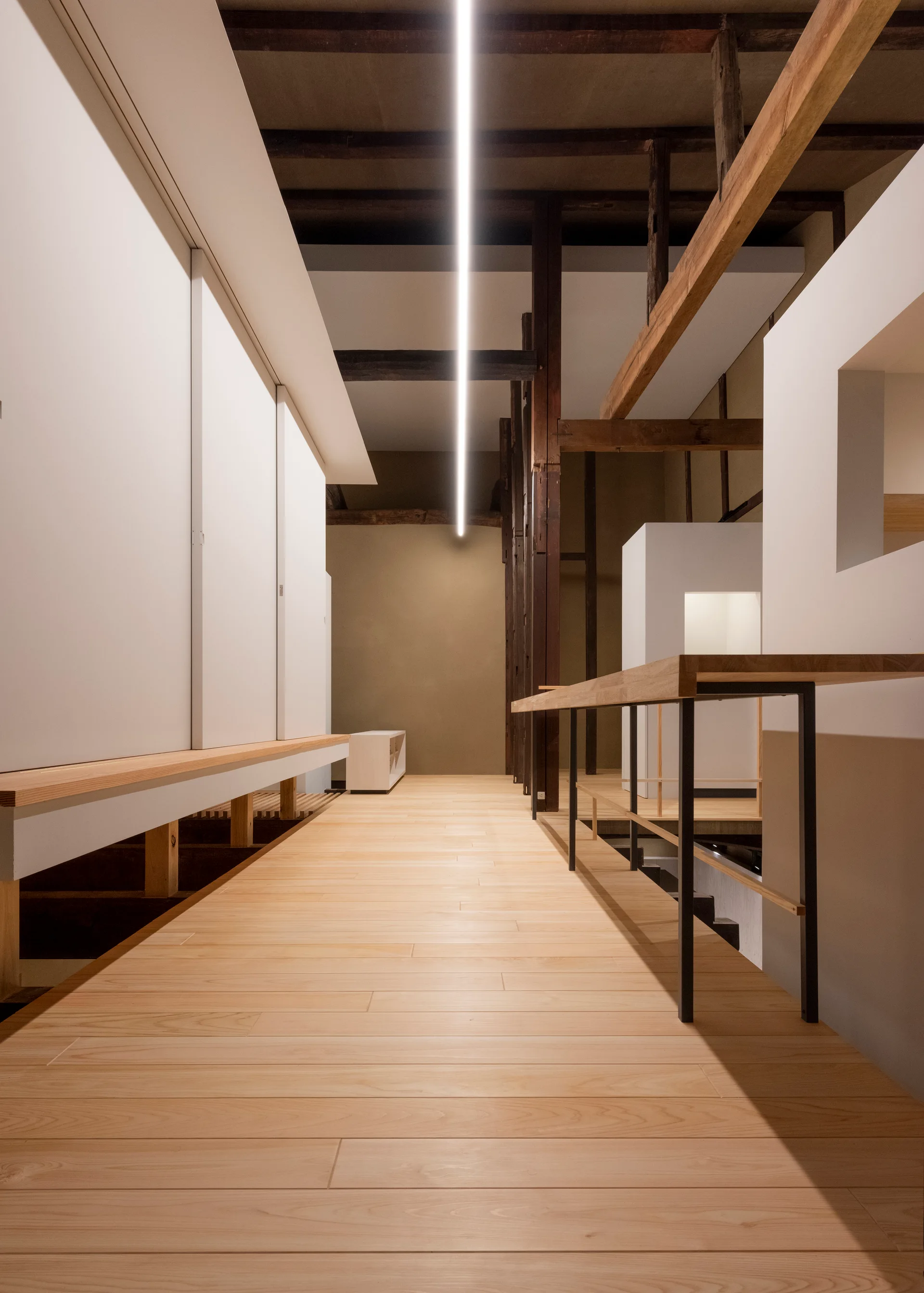 A minimalist hallway featuring light wood floors, rustic dark ceiling beams, smooth white walls, and a single long linear light strip.