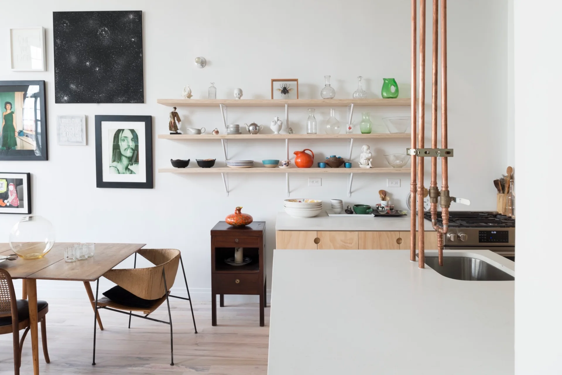 A modern kitchen and dining space features white walls, open wooden shelves, a dining table, and exposed vertical copper pipes.